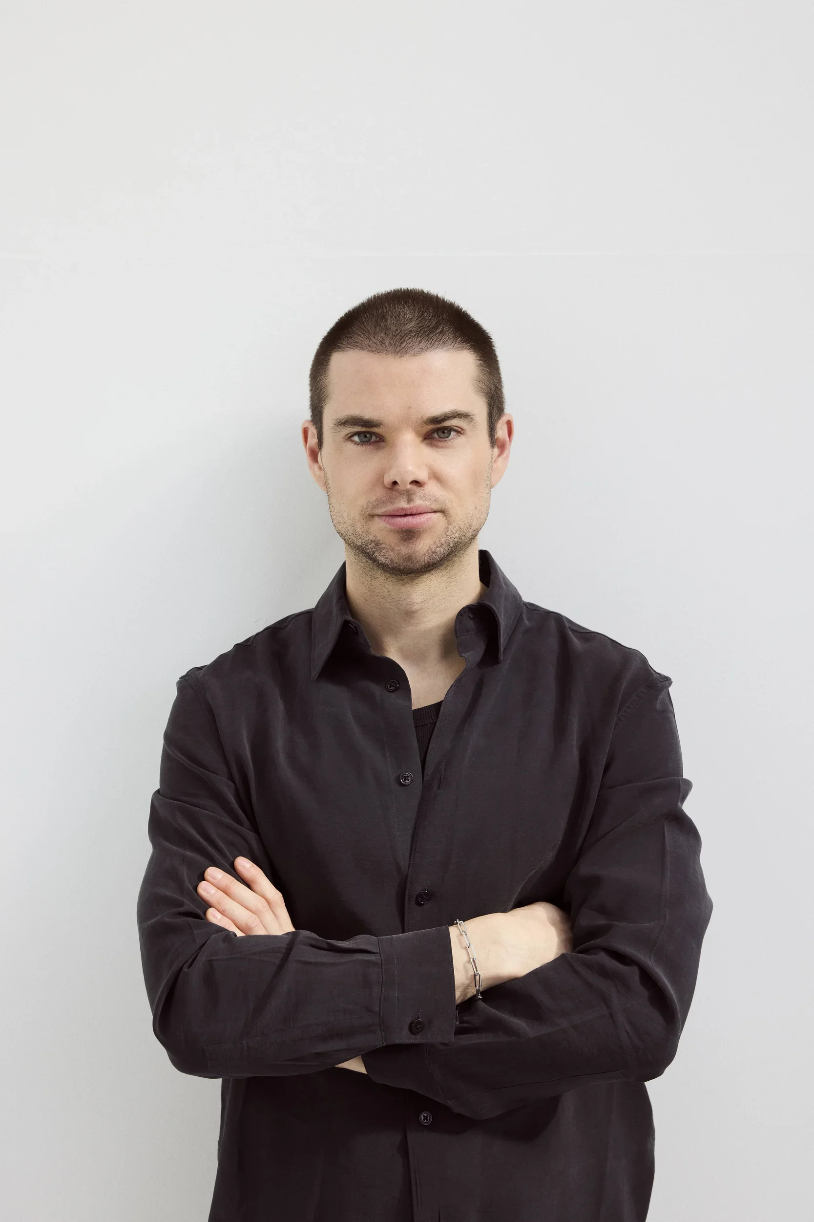 Portrait of a young man with short dark hair, dressed in a black button-up shirt, standing with arms crossed against a plain white background.