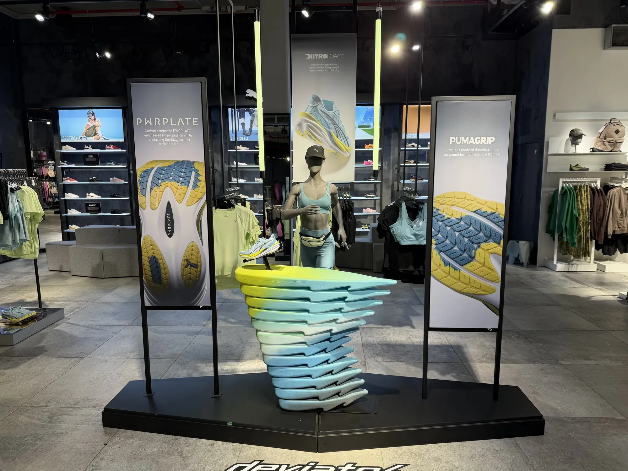 Display of Porsche and Puma branded merchandise, including clothing, shoes, and hats in a retail store, with a mannequin dressed in a black jacket and yellow pants.