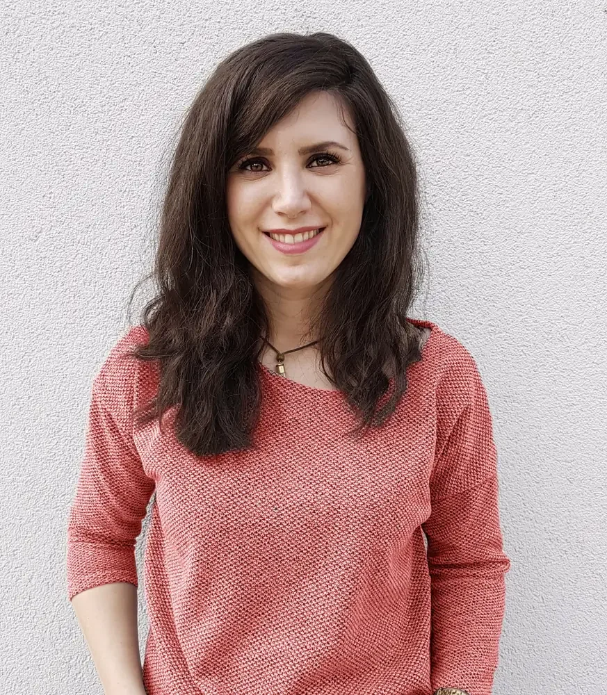 A woman with long dark hair and light skin, smiling and wearing a red textured top, standing against a plain light-colored wall.