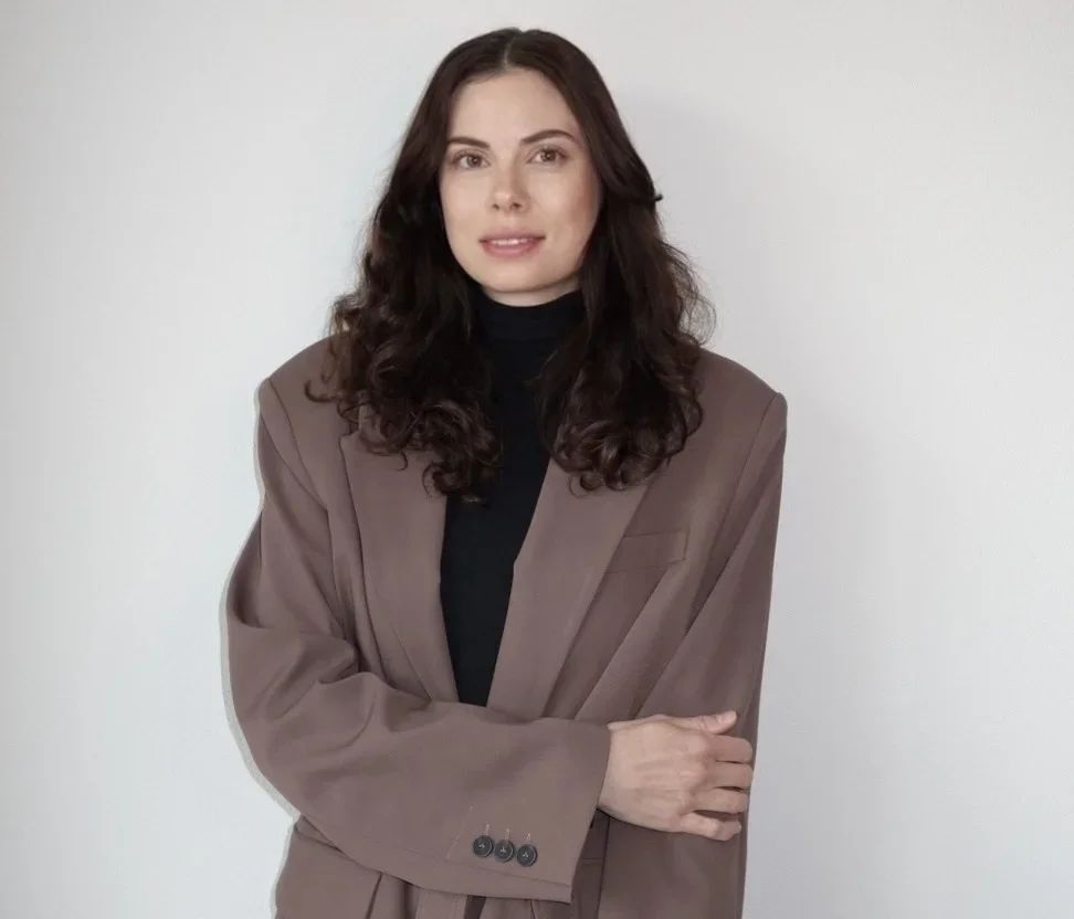 A woman with long, wavy dark hair wearing a black turtleneck and a beige blazer standing against a plain white wall.