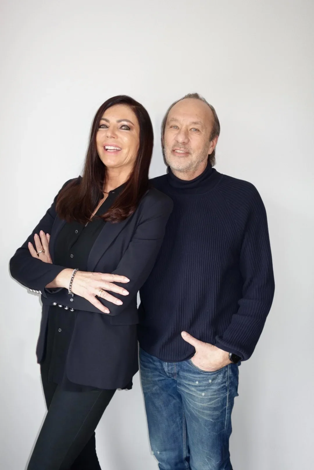 A woman with long dark hair, dressed in a black blazer and black pants, standing with arms crossed, smiling, next to a man with short hair, wearing a navy sweater and jeans, standing against a plain white background.