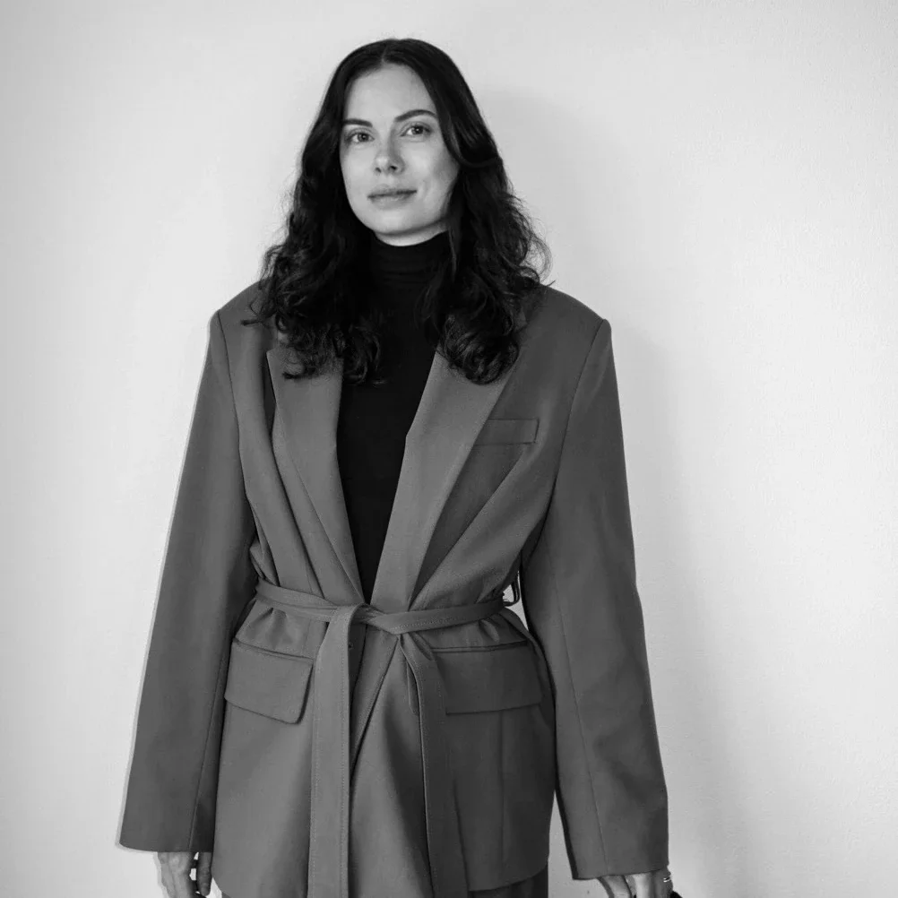 A woman with long, wavy dark hair wearing a black turtleneck and a beige blazer standing against a plain white wall.