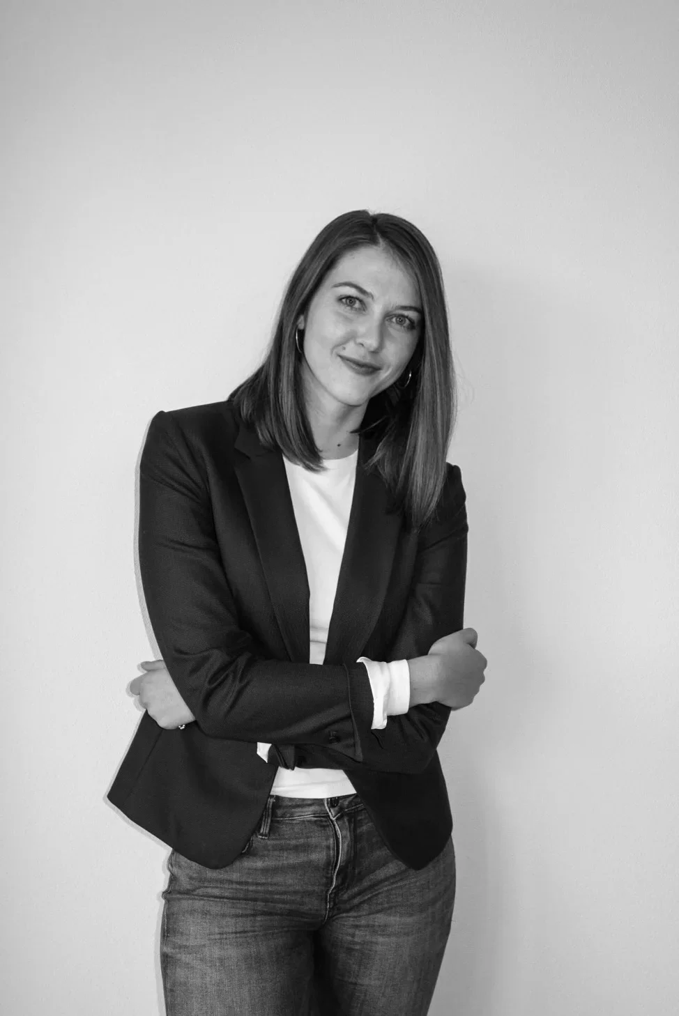 A young woman with shoulder-length brown hair, wearing a black blazer over a white top, smiling while standing against a plain white wall.