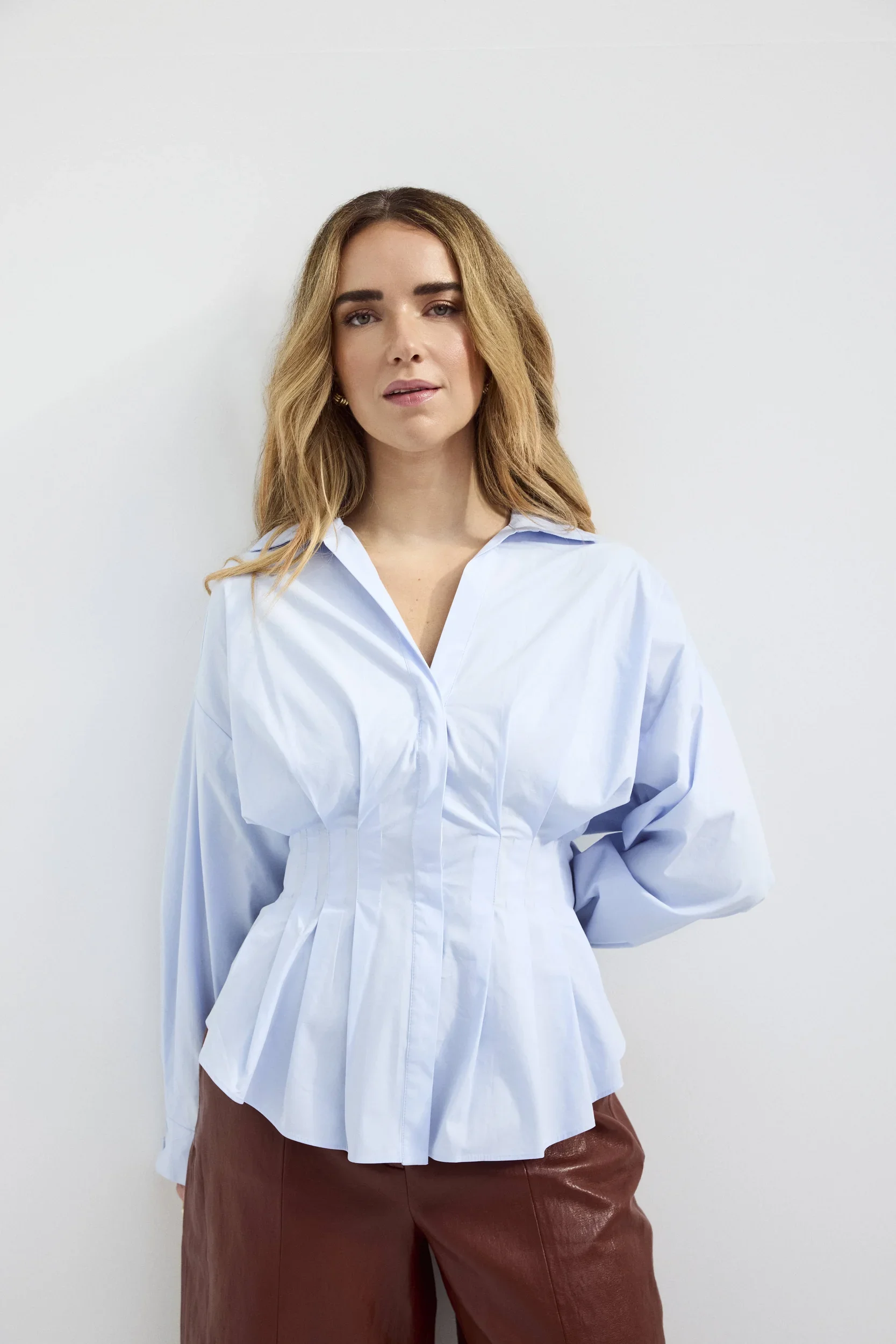 A woman with wavy blonde hair wearing a white blouse and brown pants standing against a plain white wall.