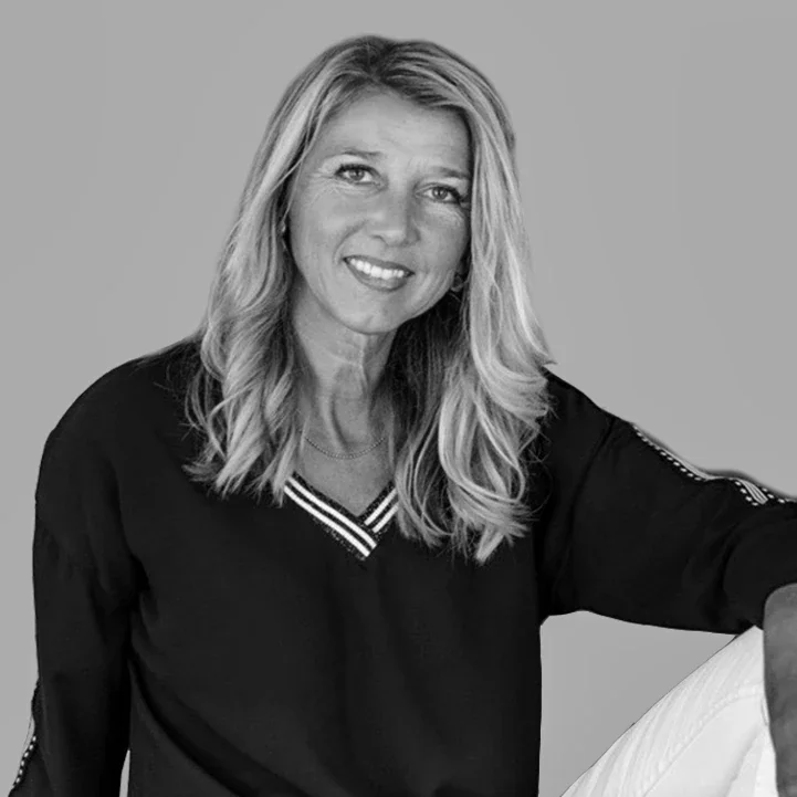 Black and white photo of a woman with long wavy hair, wearing a dark long-sleeve top with a V-neck and white pants, sitting relaxed with one arm resting on her knee, smiling at the camera.