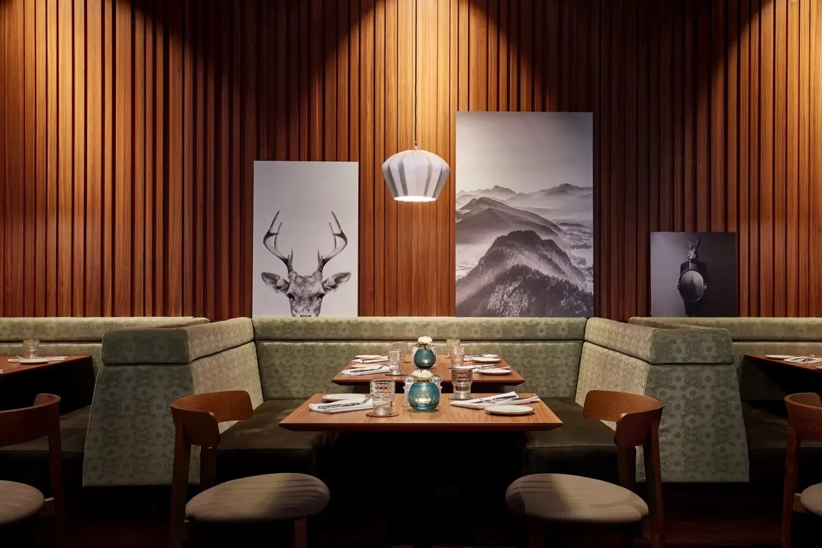 A restaurant dining area with a wooden-paneled wall adorned with art including a deer illustration, mountain landscape, and a deer with antlers holding a shield. There are tables set with glasses, plates, and cutlery, and a hanging pendant light above.