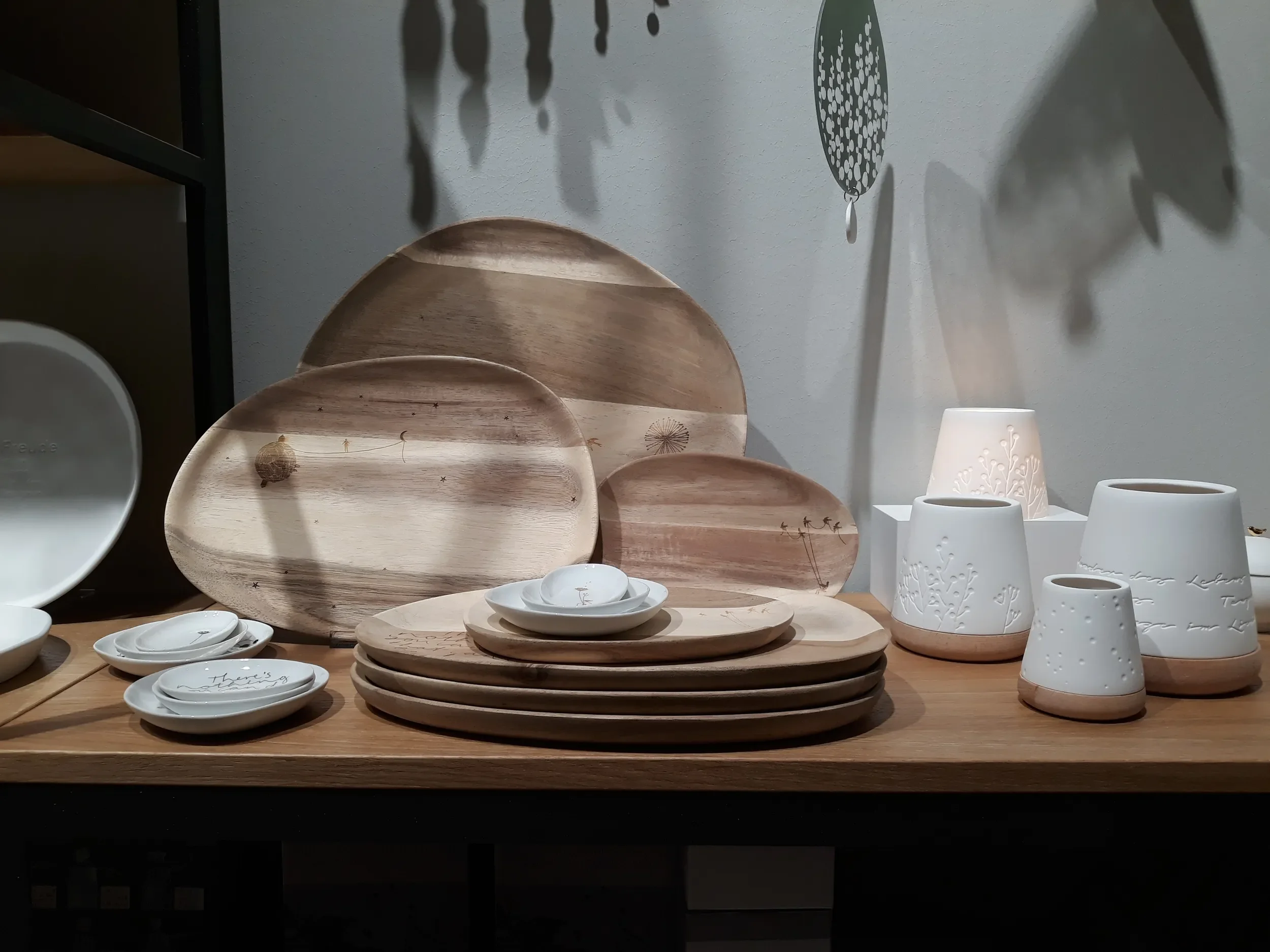 Display of white and wood-colored ceramic kitchenware including plates, bowls, and vases arranged on a wooden table with shadows cast from decorative wall hangings.