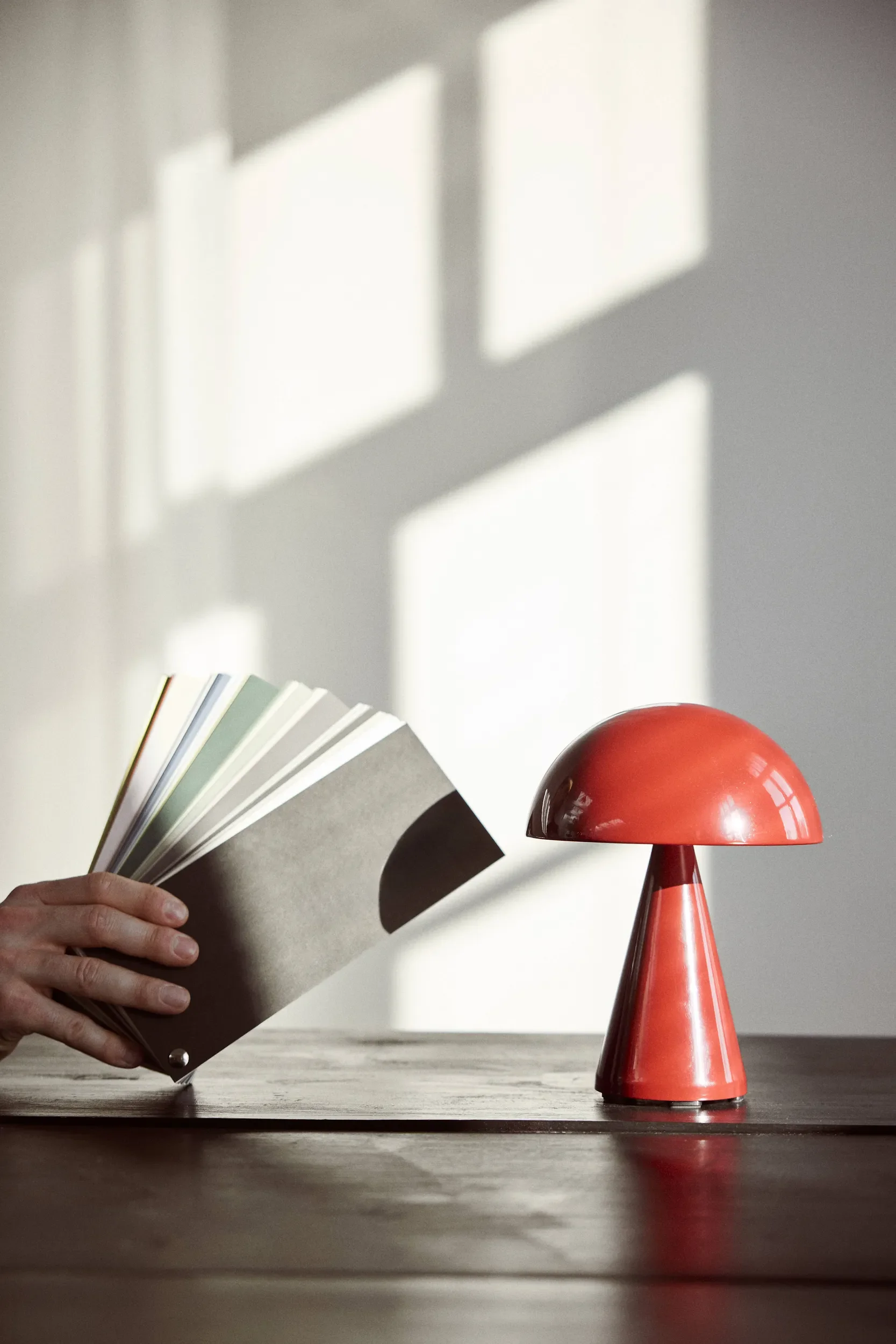 A hand holding a fan of color swatches facing a red mushroom-shaped lamp on a wooden table, with sunlight casting geometric shadows on the wall behind.