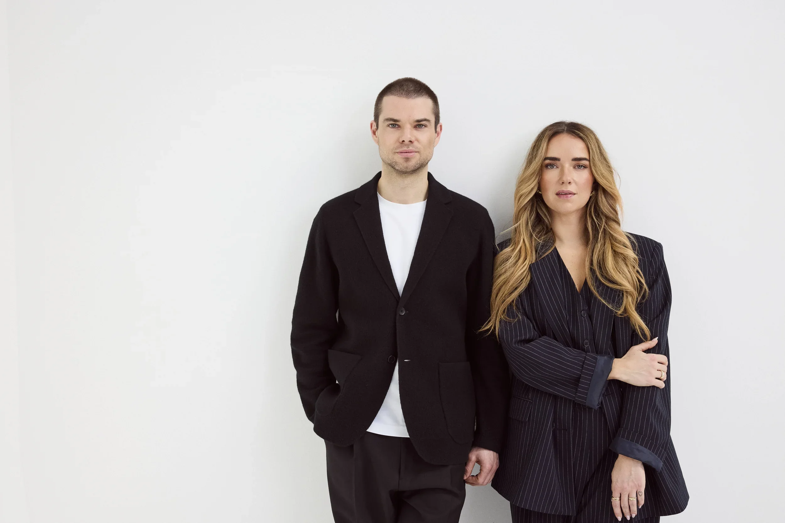 A man and woman stand side by side against a plain white wall. The man has short dark hair and is wearing a black blazer over a white shirt. The woman has long, wavy, blonde hair and is wearing a dark pinstripe blazer. Both have neutral expressions.