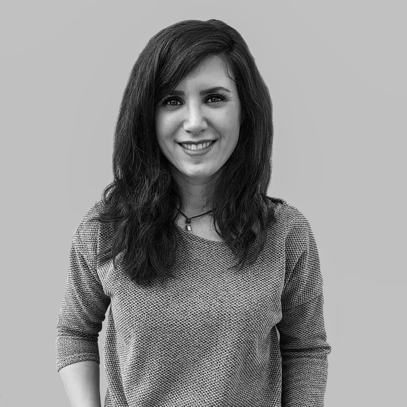 A woman with long dark hair and light skin, smiling and wearing a red textured top, standing against a plain light-colored wall.