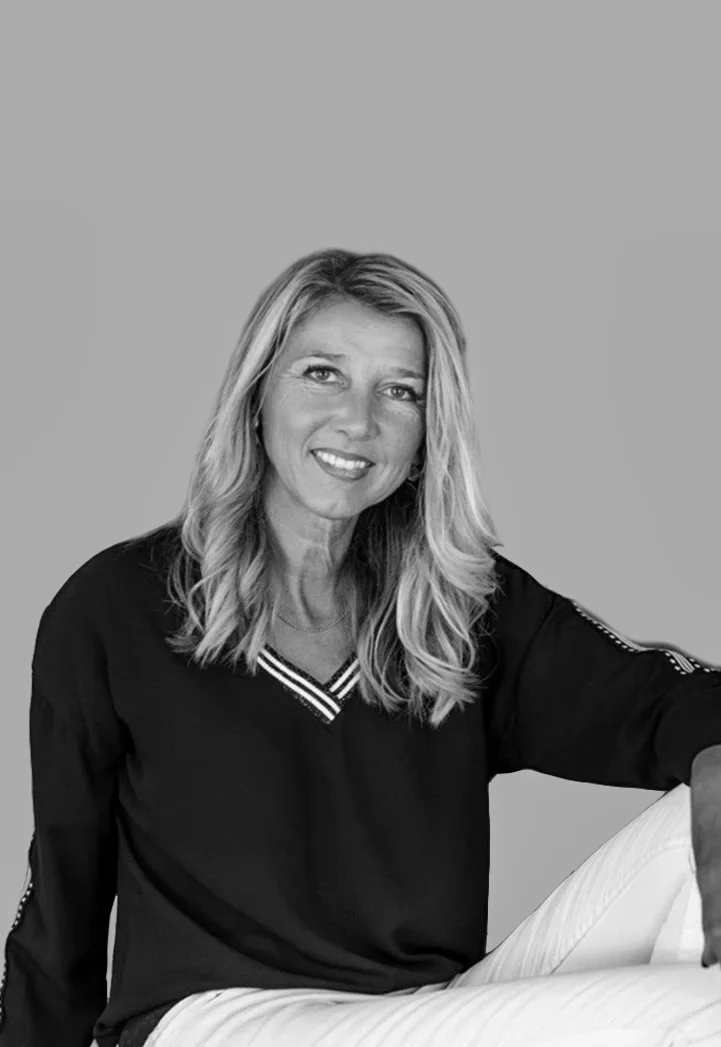 Black and white photo of a woman with long wavy hair, wearing a dark long-sleeve top with a V-neck and white pants, sitting relaxed with one arm resting on her knee, smiling at the camera.