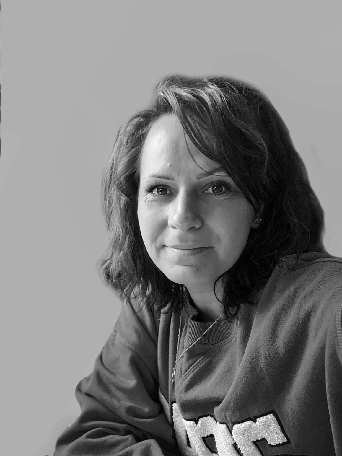 Black-and-white portrait of a woman with shoulder-length wavy hair, wearing a sweatshirt and a necklace, looking directly at the camera with a slight smile.
