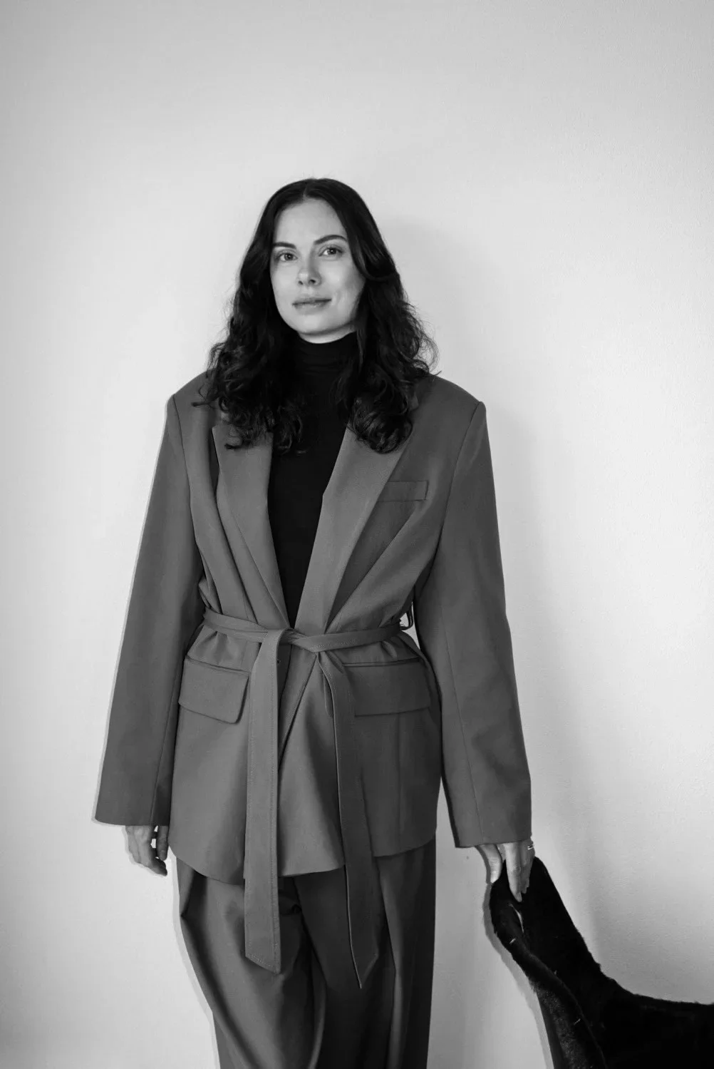 A woman with long, wavy dark hair wearing a black turtleneck and a beige blazer standing against a plain white wall.