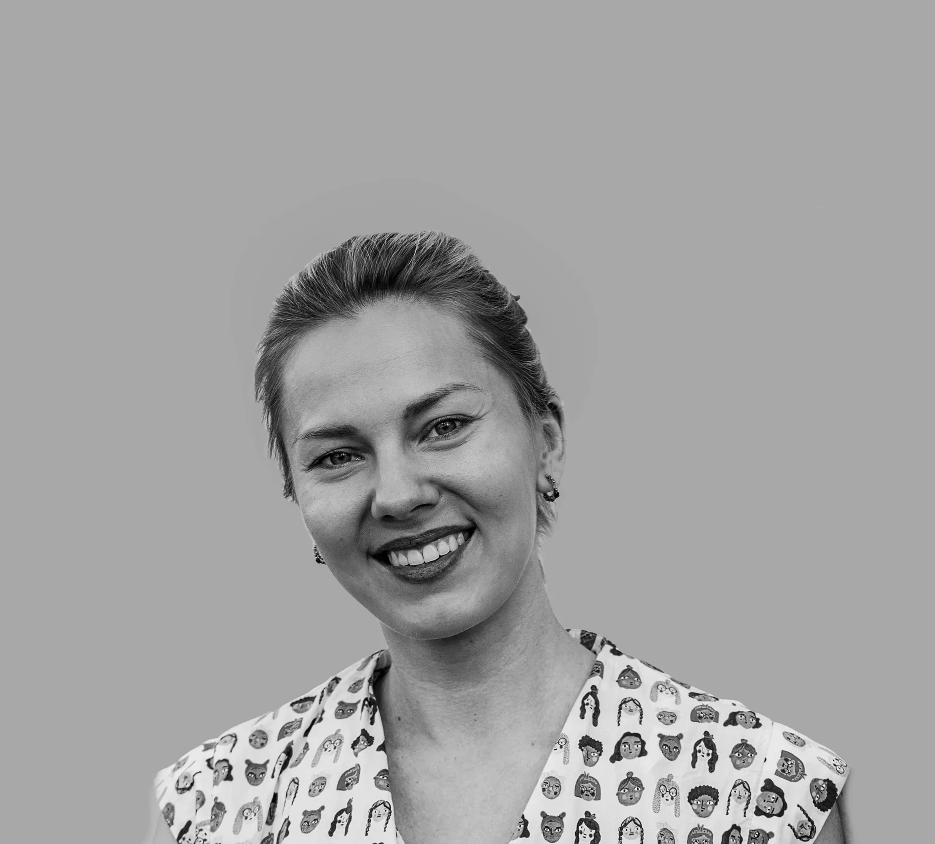 Black and white portrait of a smiling woman with light-colored hair tied back, wearing a shirt with cartoon face illustrations.