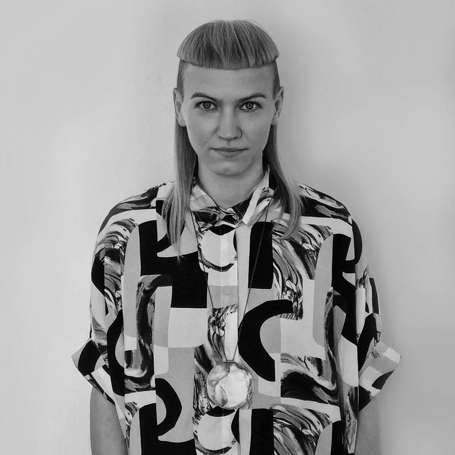 A person with Blonde hair in a Madonna style haircut, wearing a colorful abstract patterned shirt with a large gold medal around their neck, standing against a plain white background.