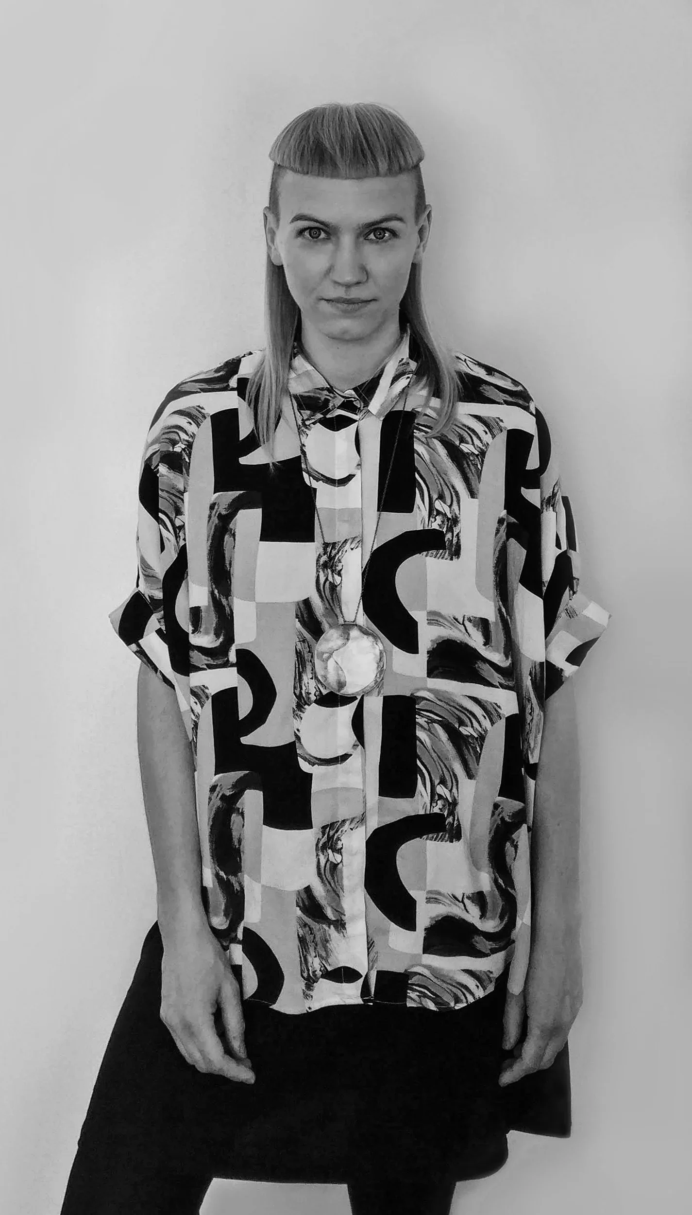 A person with Blonde hair in a Madonna style haircut, wearing a colorful abstract patterned shirt with a large gold medal around their neck, standing against a plain white background.