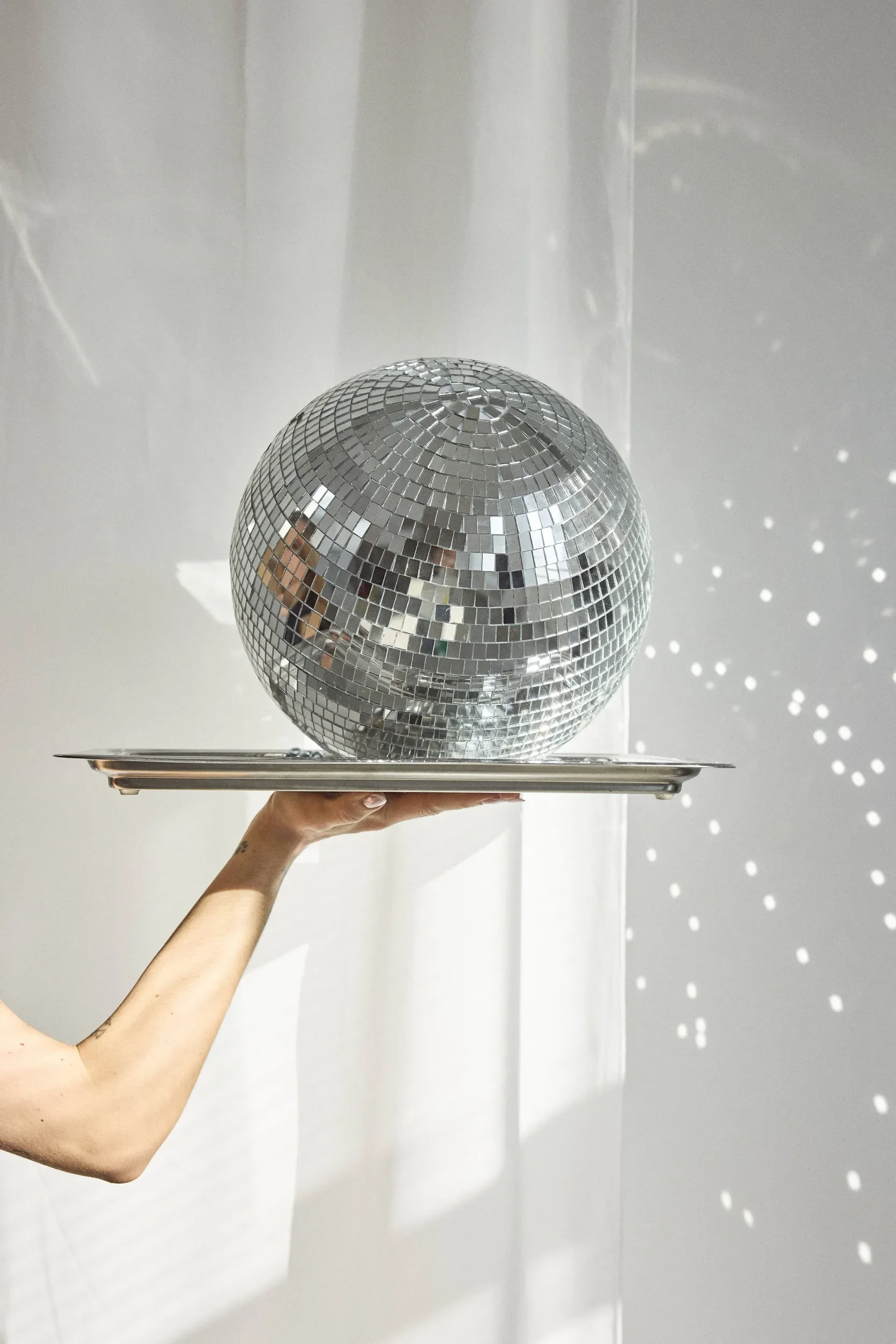 A disco ball resting on a silver tray, held by a person's hand with a minimal background and light reflections.