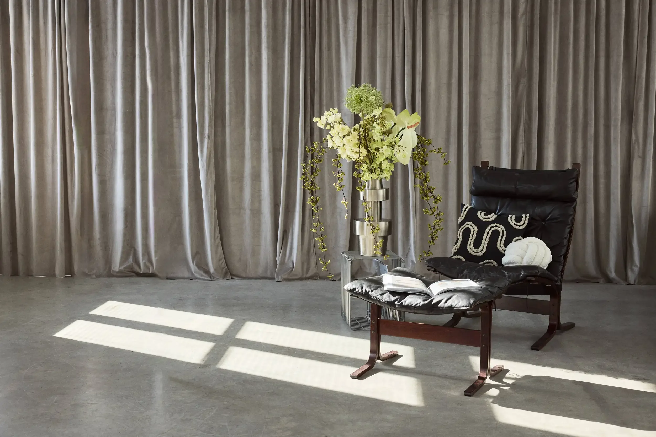 A modern interior with a black leather lounge chair, an open book on a matching ottoman, a large silver vase with white flowers and trailing greenery, grey curtains in the background, and shadows of window panes cast on the floor.