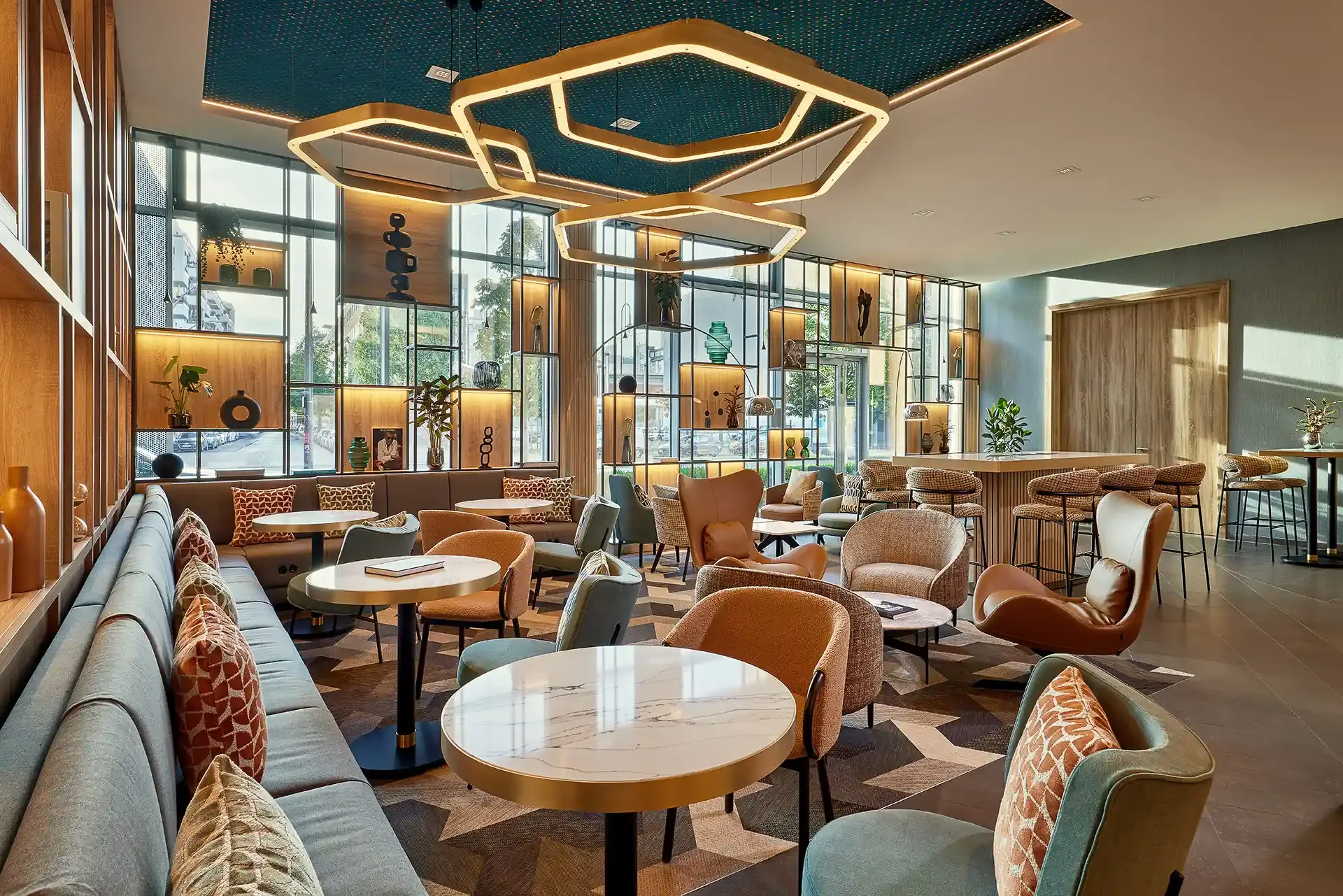 Modern hotel lobby with large windows, decorative shelving displaying vases and art pieces, and stylish seating including armchairs and bar stools. The room features warm wood accents, geometric ceiling light fixtures, and a patterned rug.