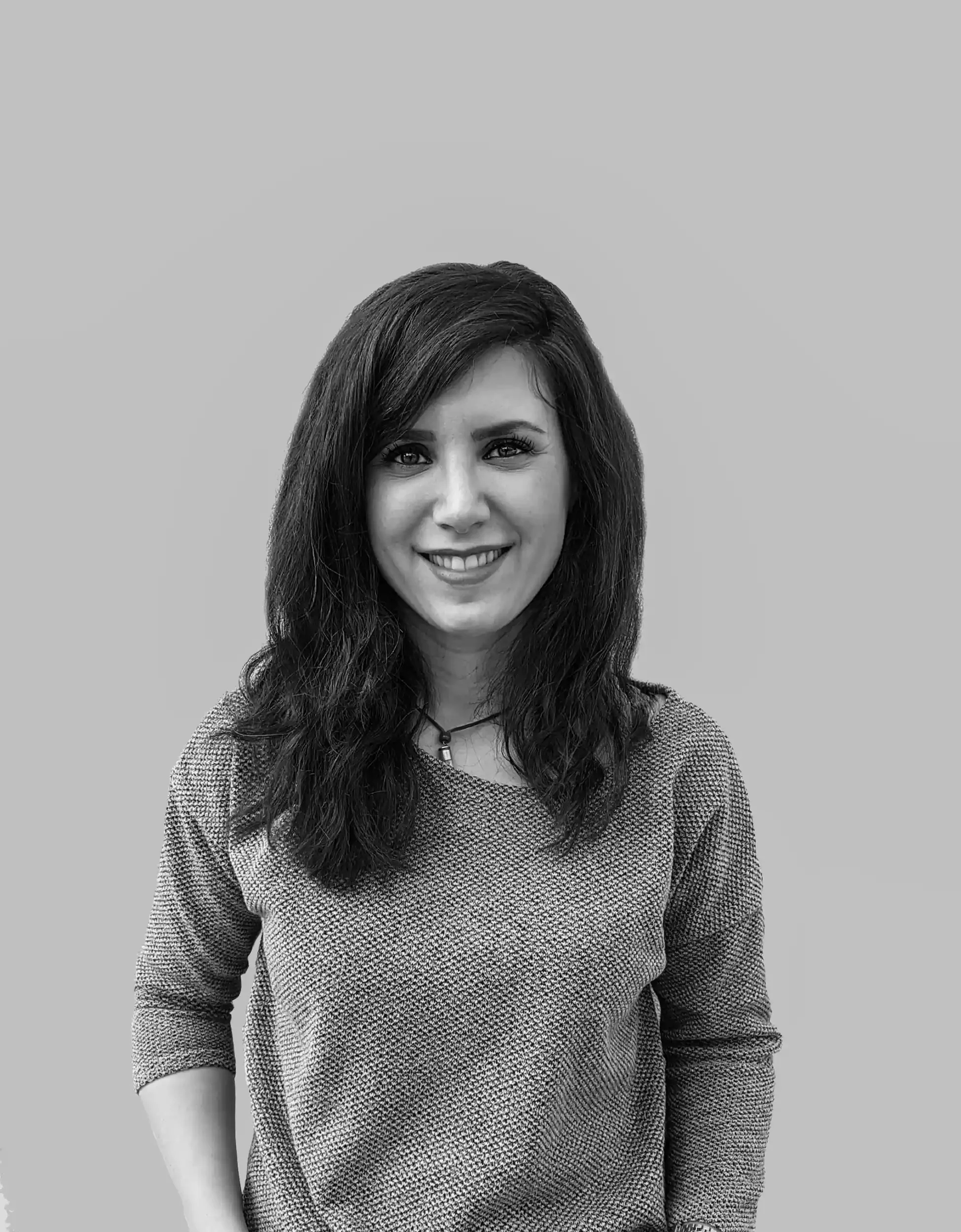 A woman with long dark hair and light skin, smiling and wearing a red textured top, standing against a plain light-colored wall.