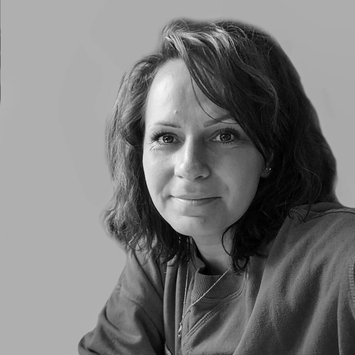 Black-and-white portrait of a woman with shoulder-length wavy hair, wearing a sweatshirt and a necklace, looking directly at the camera with a slight smile.