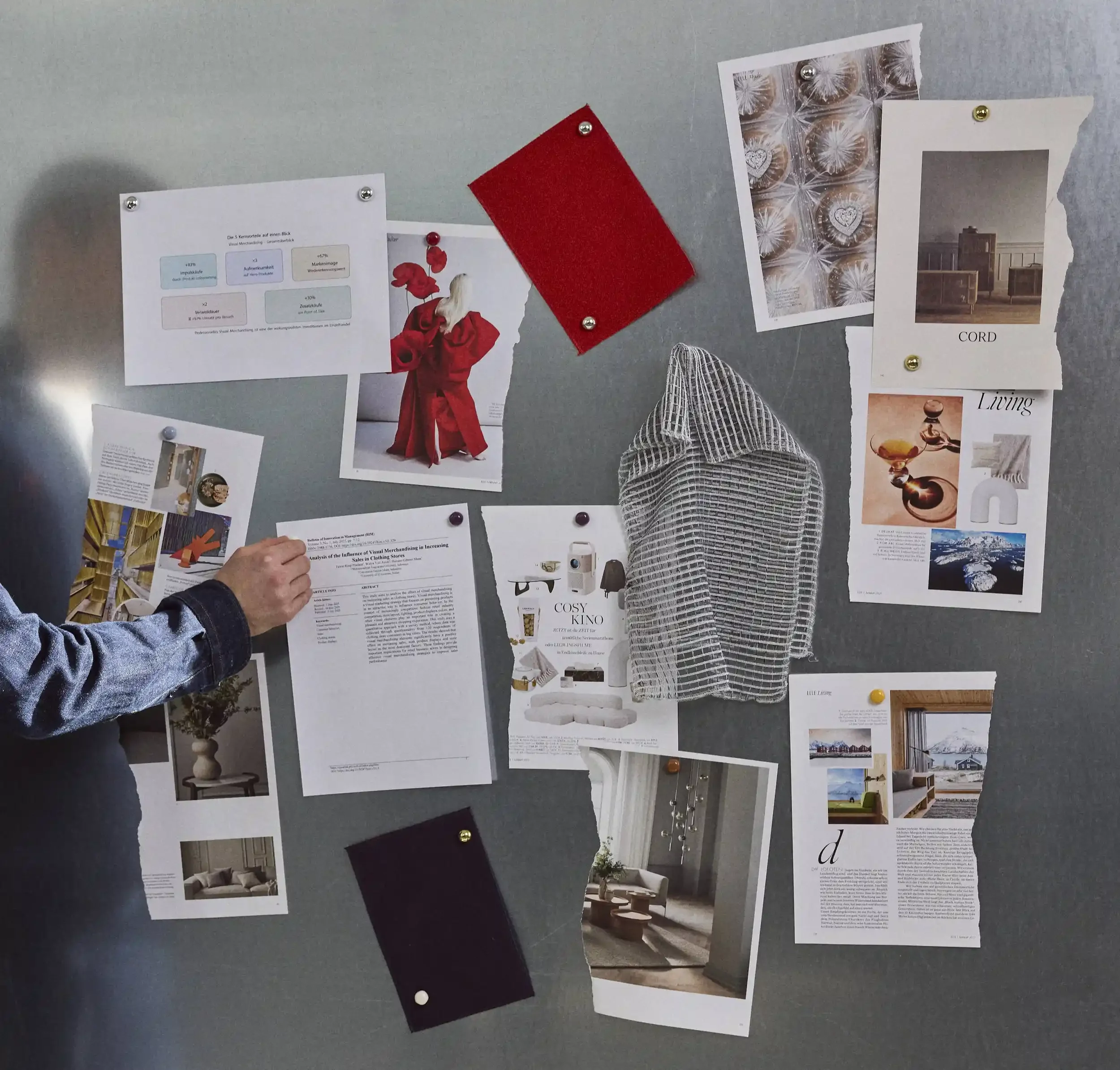 A gray bulletin board with various magazine clippings, photographs, and color swatches pinned with small decorative pushpins. The images depict interior design, decor, and products, with a person's hand reaching towards one of the papers.