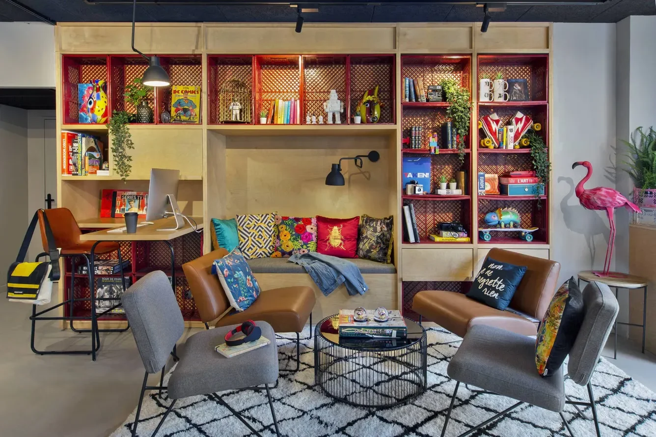 Colorful living room with wall-to-wall shelving, seating area with chairs and a sofa, decorative pillows, bookshelf filled with books and decor, pink flamingo statue, and a patterned rug.