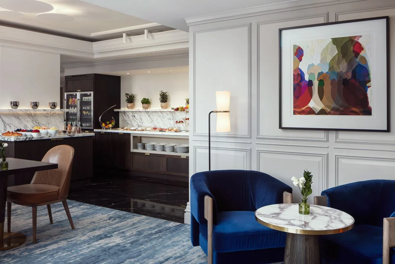Elegant hotel lounge with dark blue velvet chairs, a marble-topped round table with a small vase of white flowers, and a white paneled wall with colorful abstract artwork. In the background, a breakfast buffet area with white marble countertops, dark cabinets, and plates stacked on shelves.
