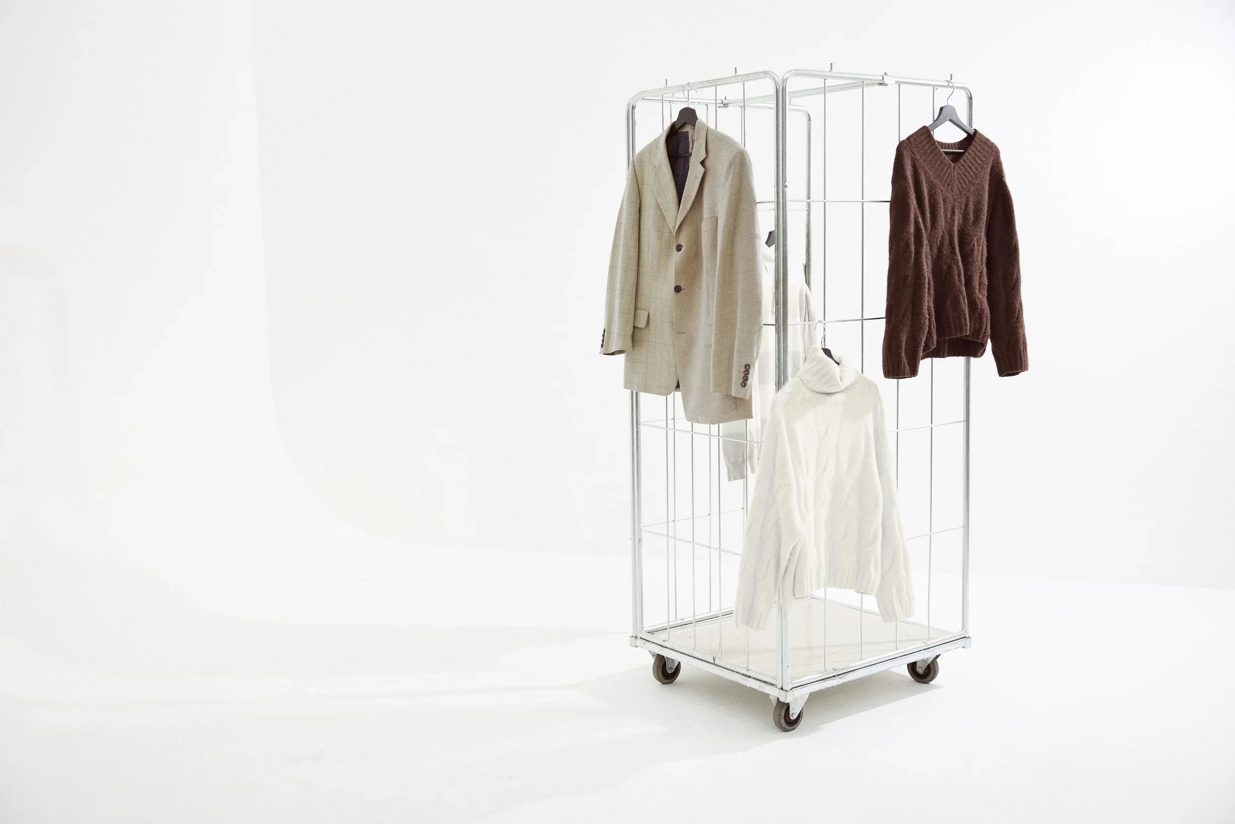 Clothes on a metal rolling rack against a white background, including a beige blazer, a brown sweater, and a white cable knit sweater.