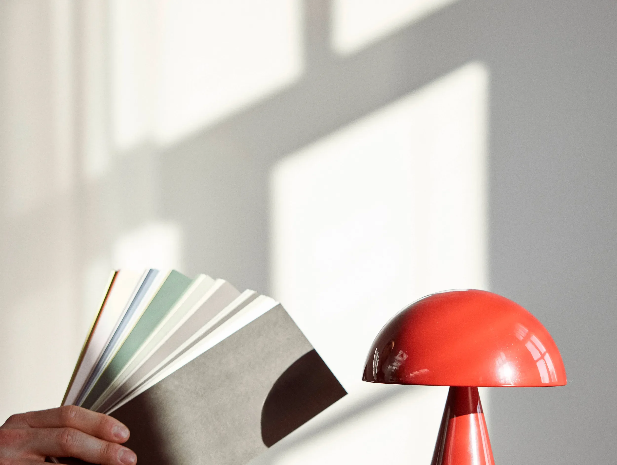 A person's hand holding a fan of color swatches near a red modern table lamp with a rounded dome-shaped shade, against a plain wall with sunlight and shadow patterns.