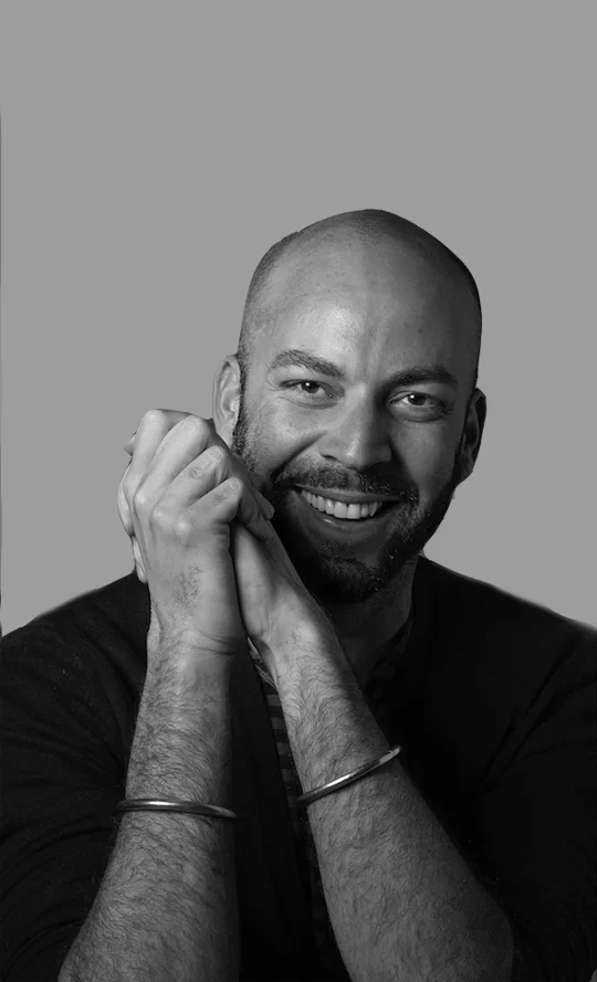 Black and white portrait of a smiling bald man with a beard, resting his chin on his hands, wearing a dark shirt and bracelets on his wrists.