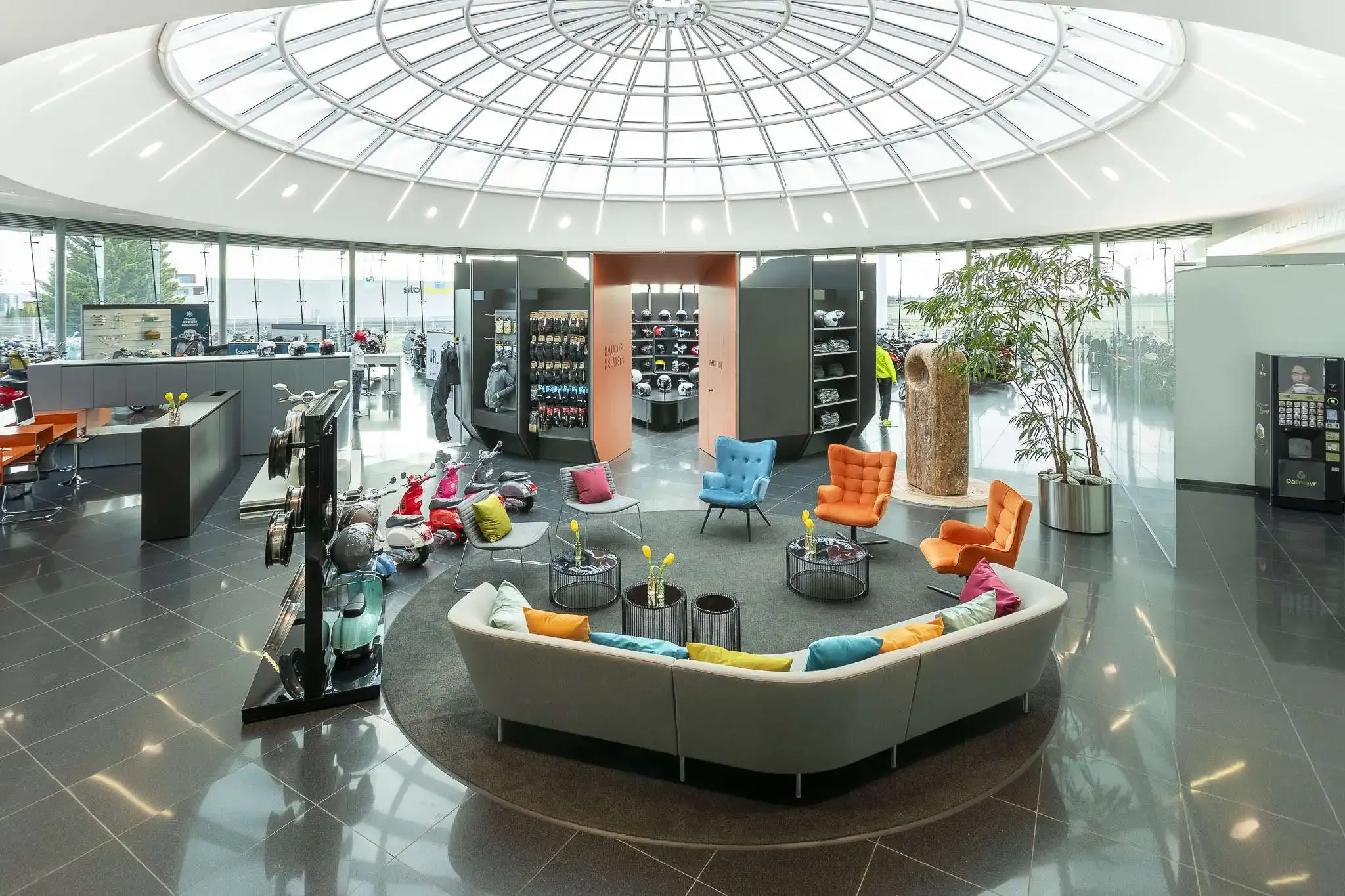 Modern indoor showroom and lounge with large glass windows, colorful chairs arranged in a circle on a rug, decorative vases with artificial flowers, display shelves with helmets and accessories, model motorcycles, potted plants, a reception desk, and a vending machine, all under a circular skylight ceiling.