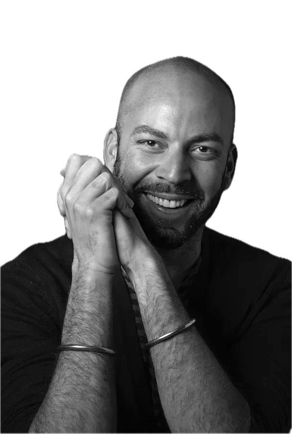 Black and white portrait of a smiling bald man with a beard, resting his chin on his hands, wearing a dark shirt and bracelets on his wrists.