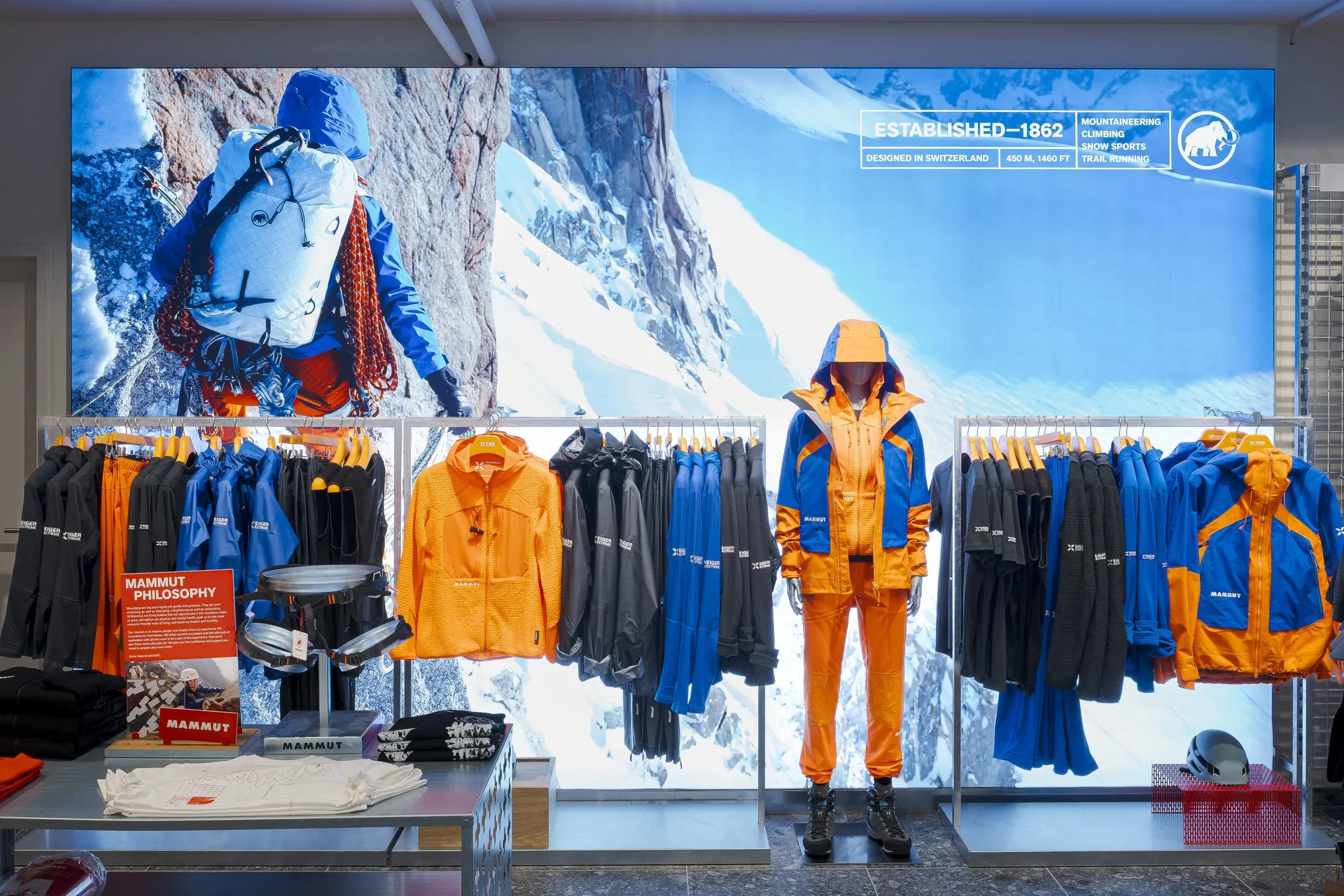 Display table with folded outdoor clothing and accessories, including jackets, pants, hats, and backpacks in a retail store.