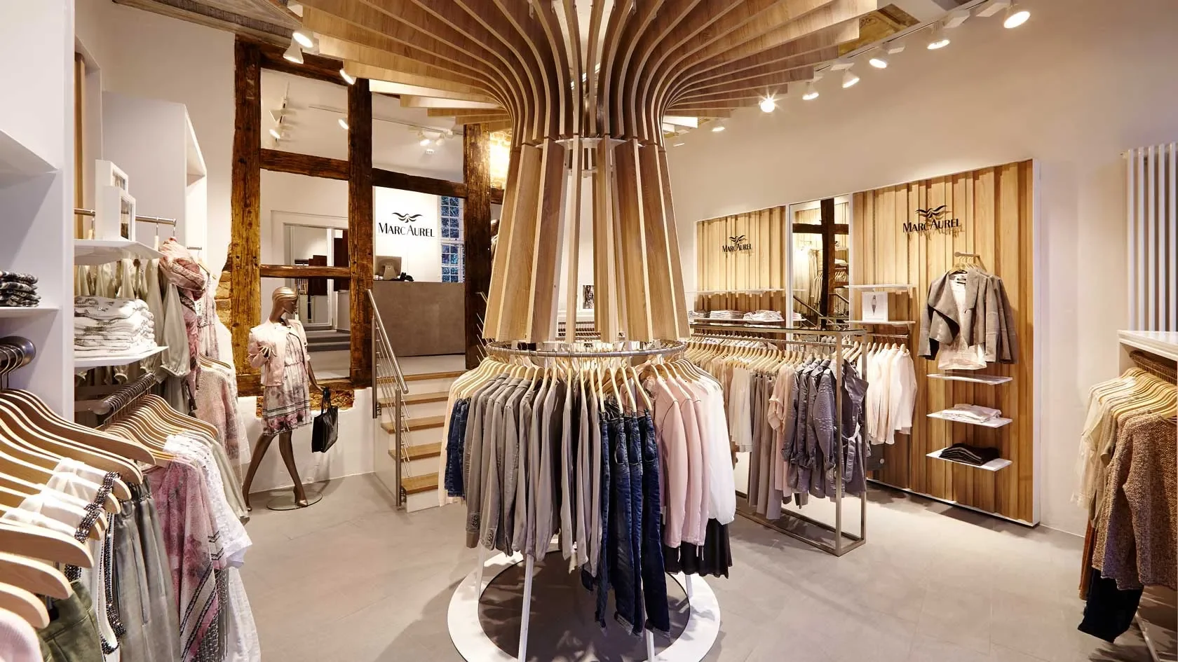 Interior view of a clothing store with circular clothing rack and mannequins, wooden accents, and the brand 'Marc Aurel' visible in the background.