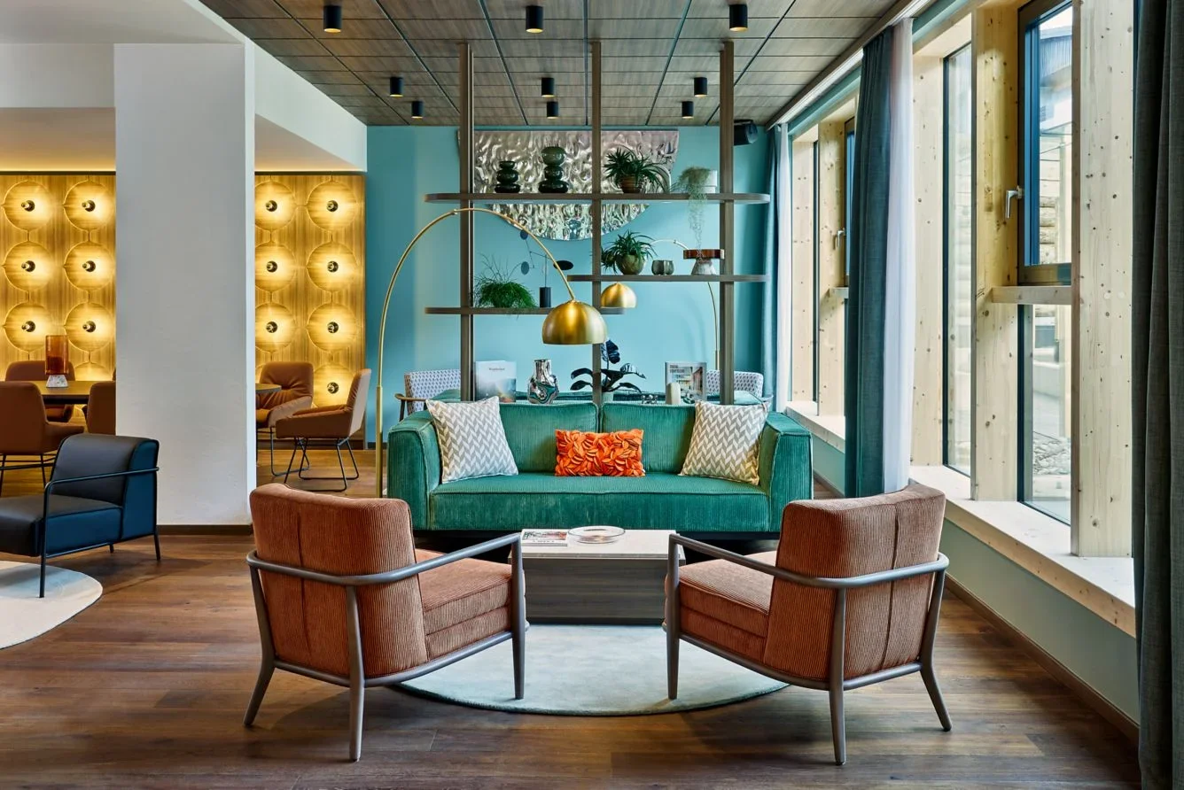 Modern hotel lobby with green sofa, brown armchairs, and large windows, decorated with plants and gold accents.