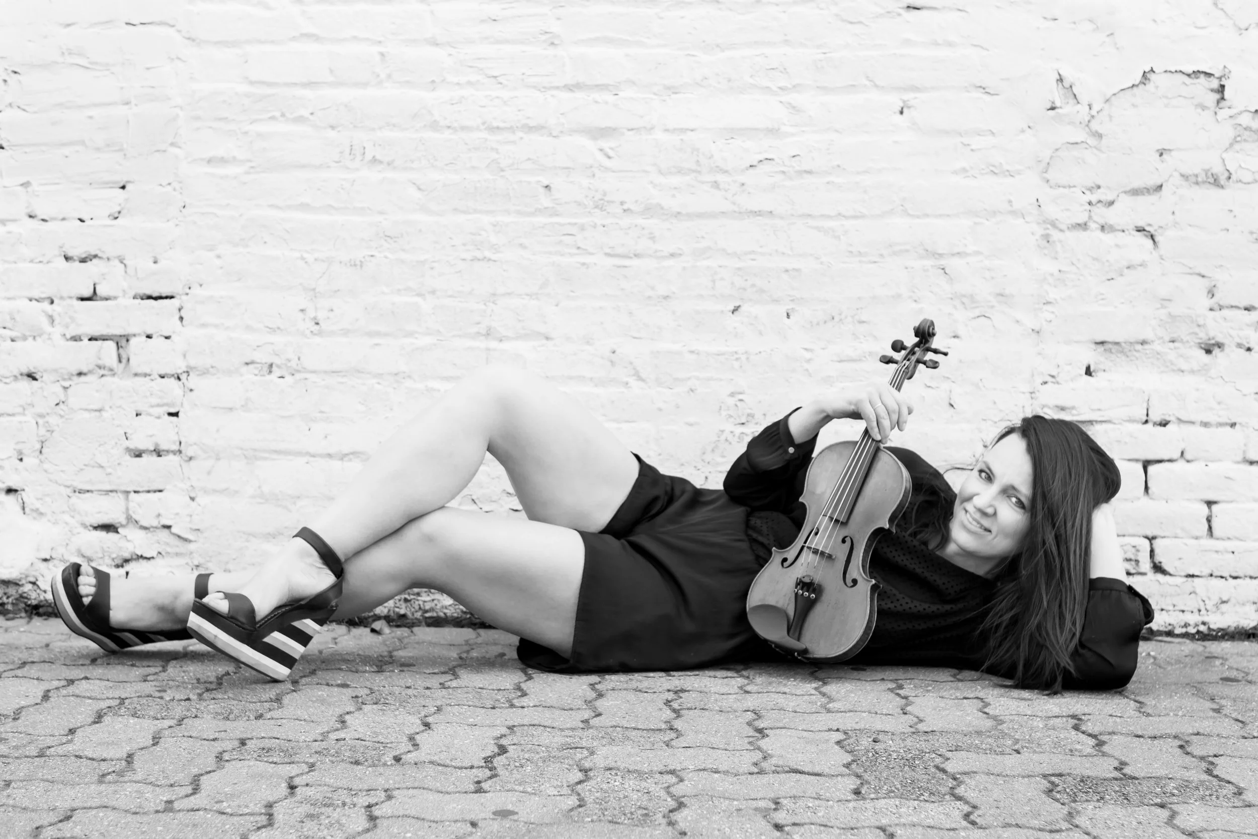 A woman lies on the ground against a brick wall, holding a violin, smiling, and wearing platform sandals and a dark dress.