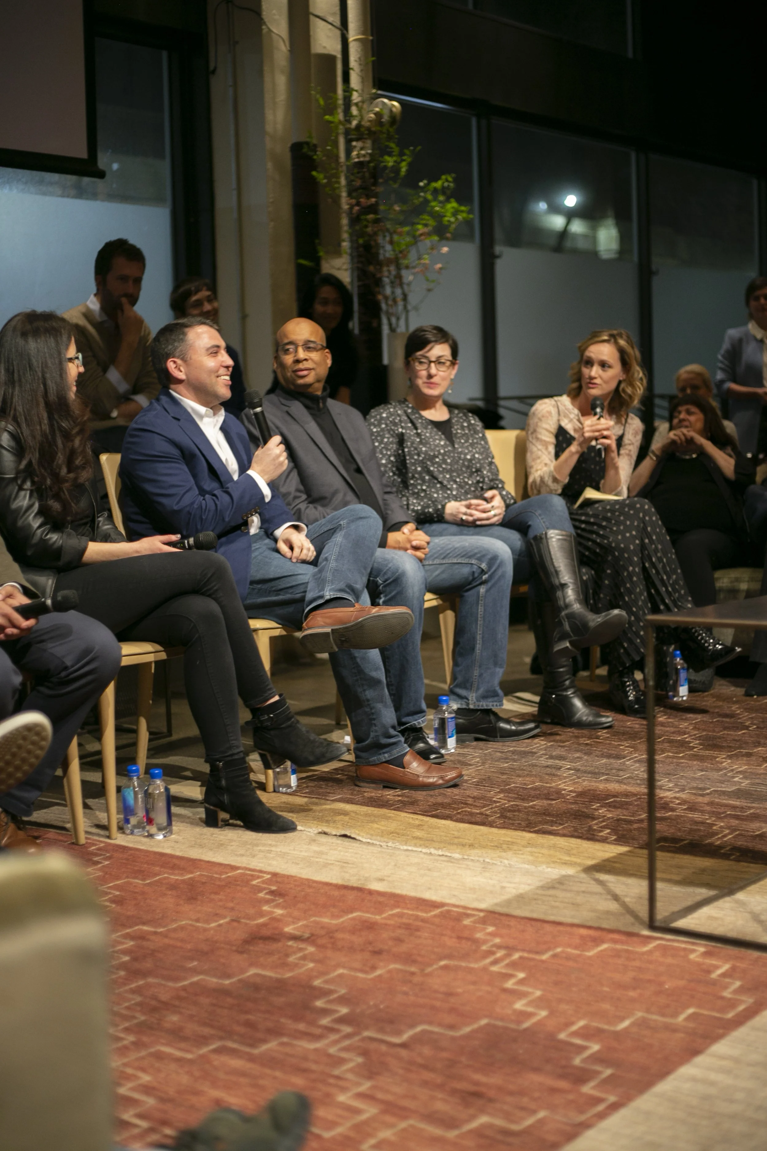 A panel discussion with five people sitting on a stage, some holding microphones, in an indoor venue with large windows and an audience in the background.