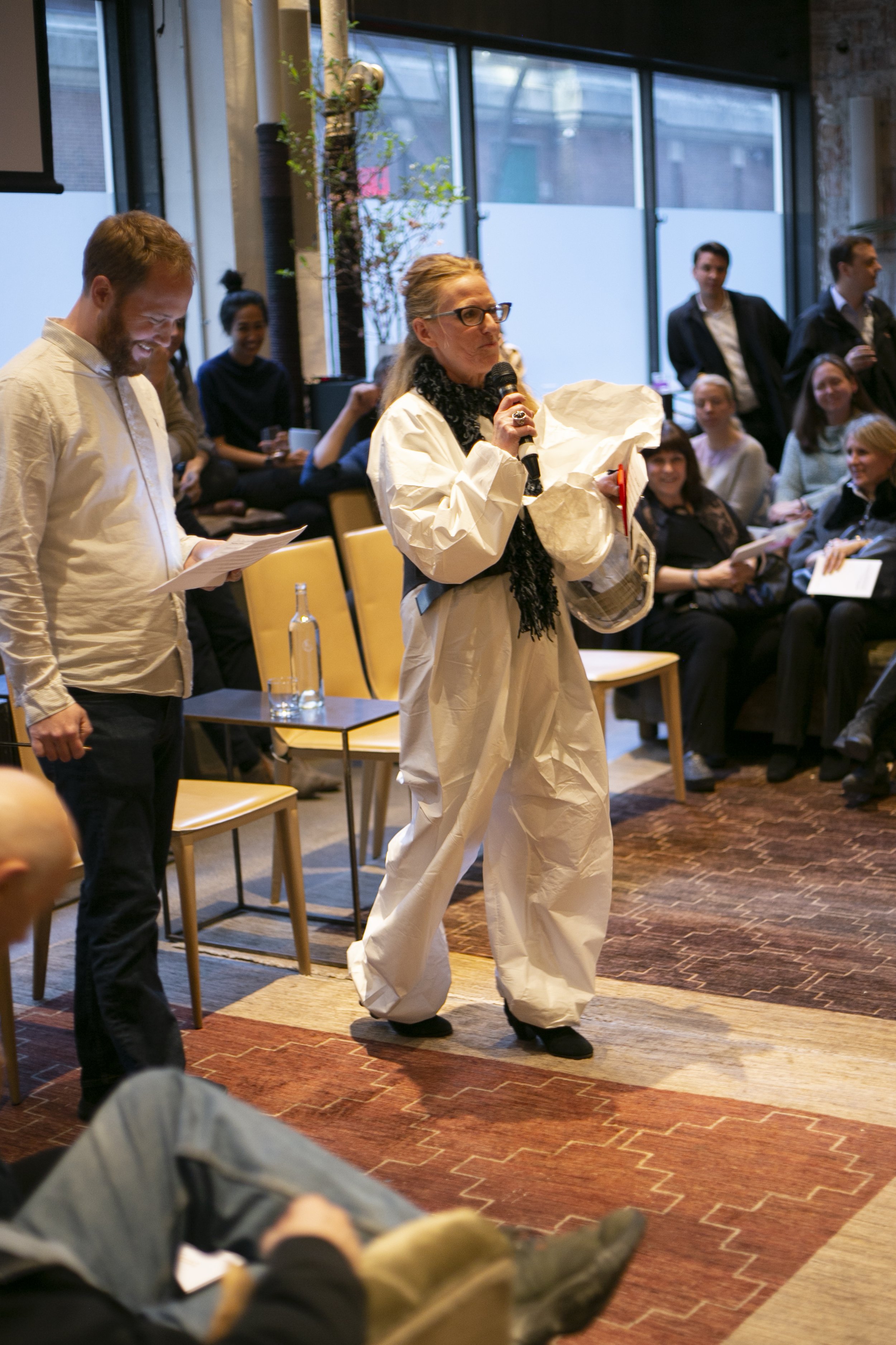 Woman speaking into a microphone at an indoor event with audience members sitting and standing around, some holding papers, in a modern, well-lit room with large windows.