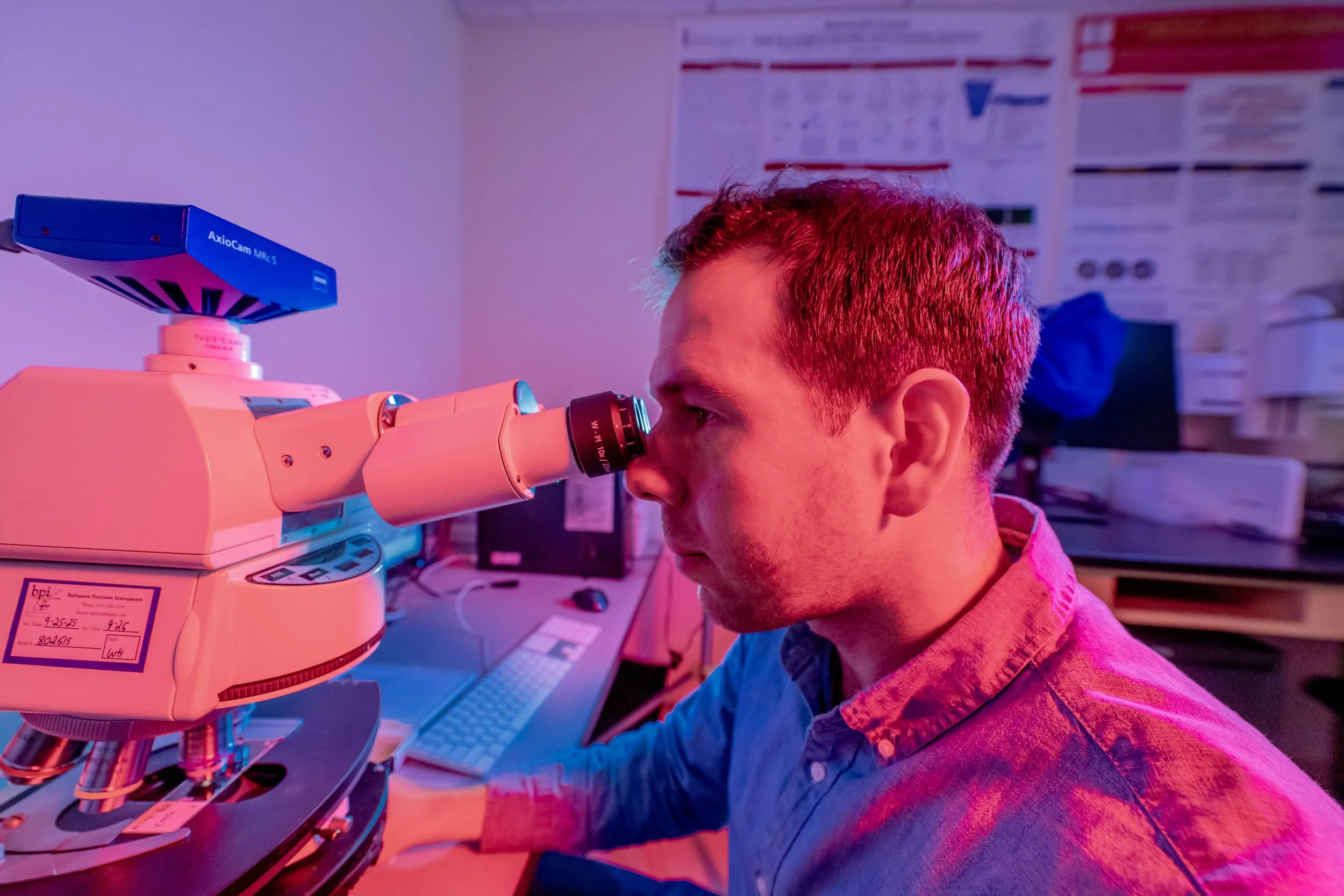 A scientist looking through a microscope in a laboratory with posters on the wall and computer equipment in the background.