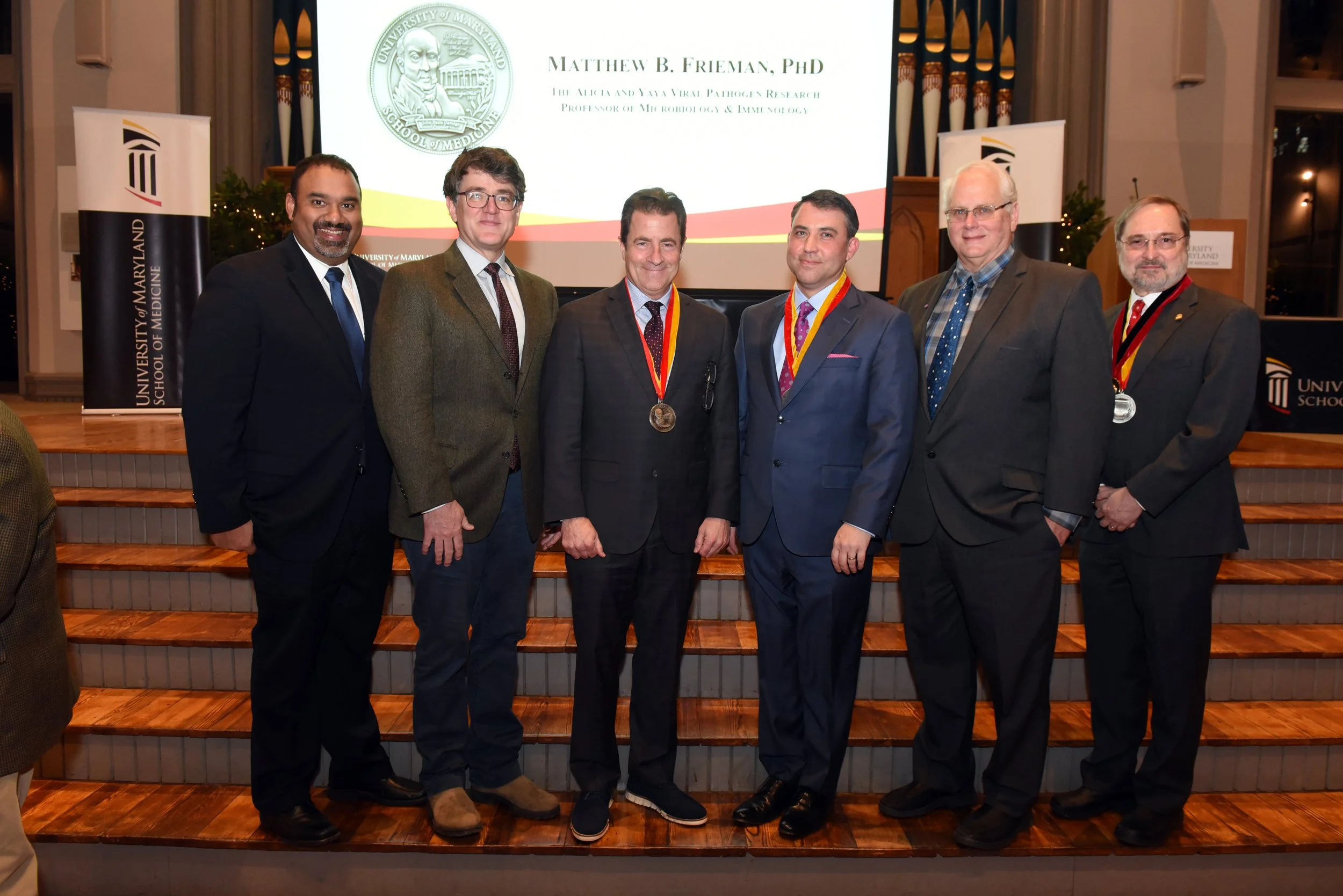 Group of seven men standing on stage in formal suits, some wearing medals around their necks, in front of a screen with the University of Maryland School of Medicine logo and the name Matthew B. Frieman, PhD. Behind them are stained glass windows and