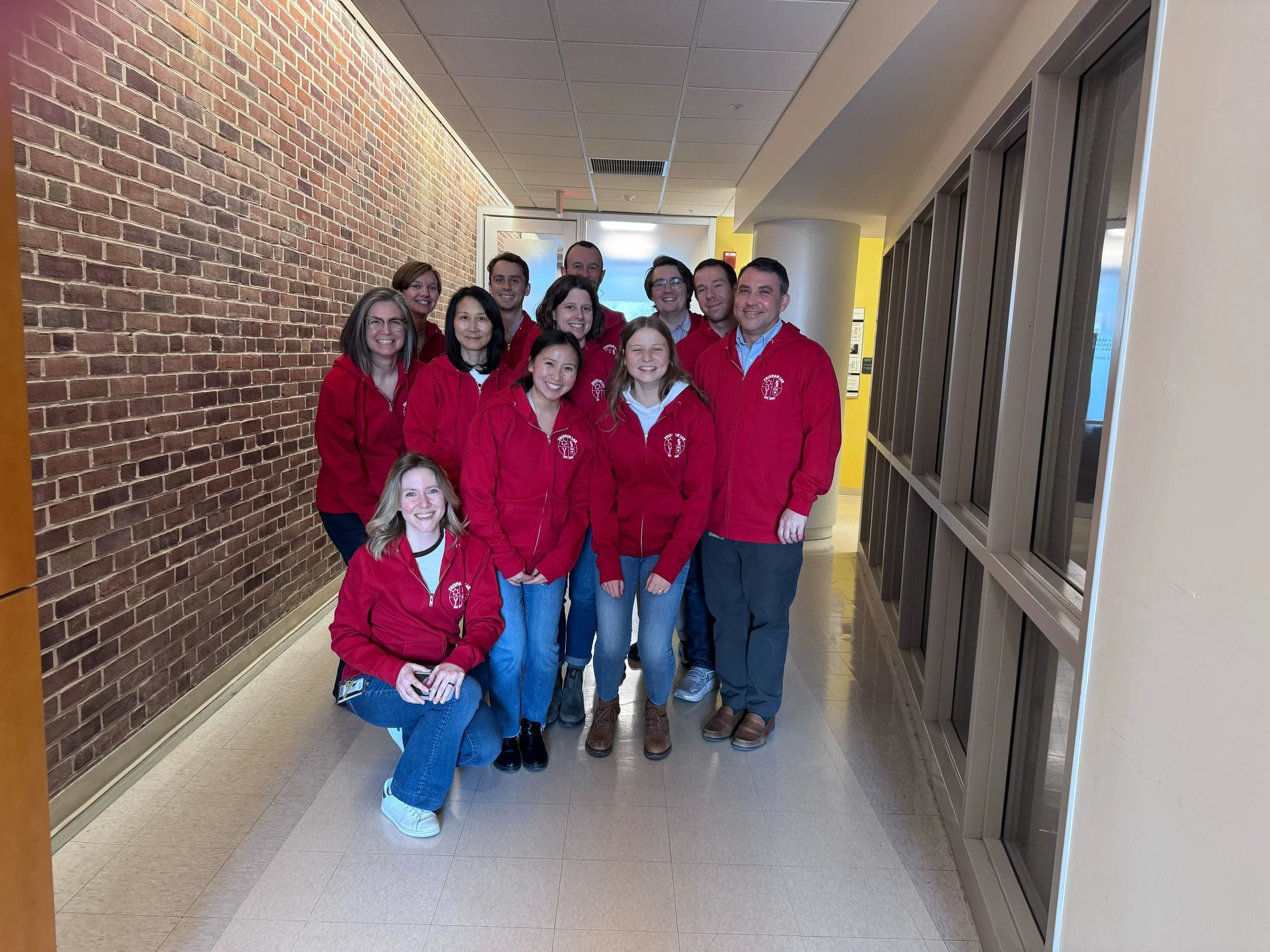 Group of people wearing red jackets standing in a hallway with brick wall on the left and windows on the right.