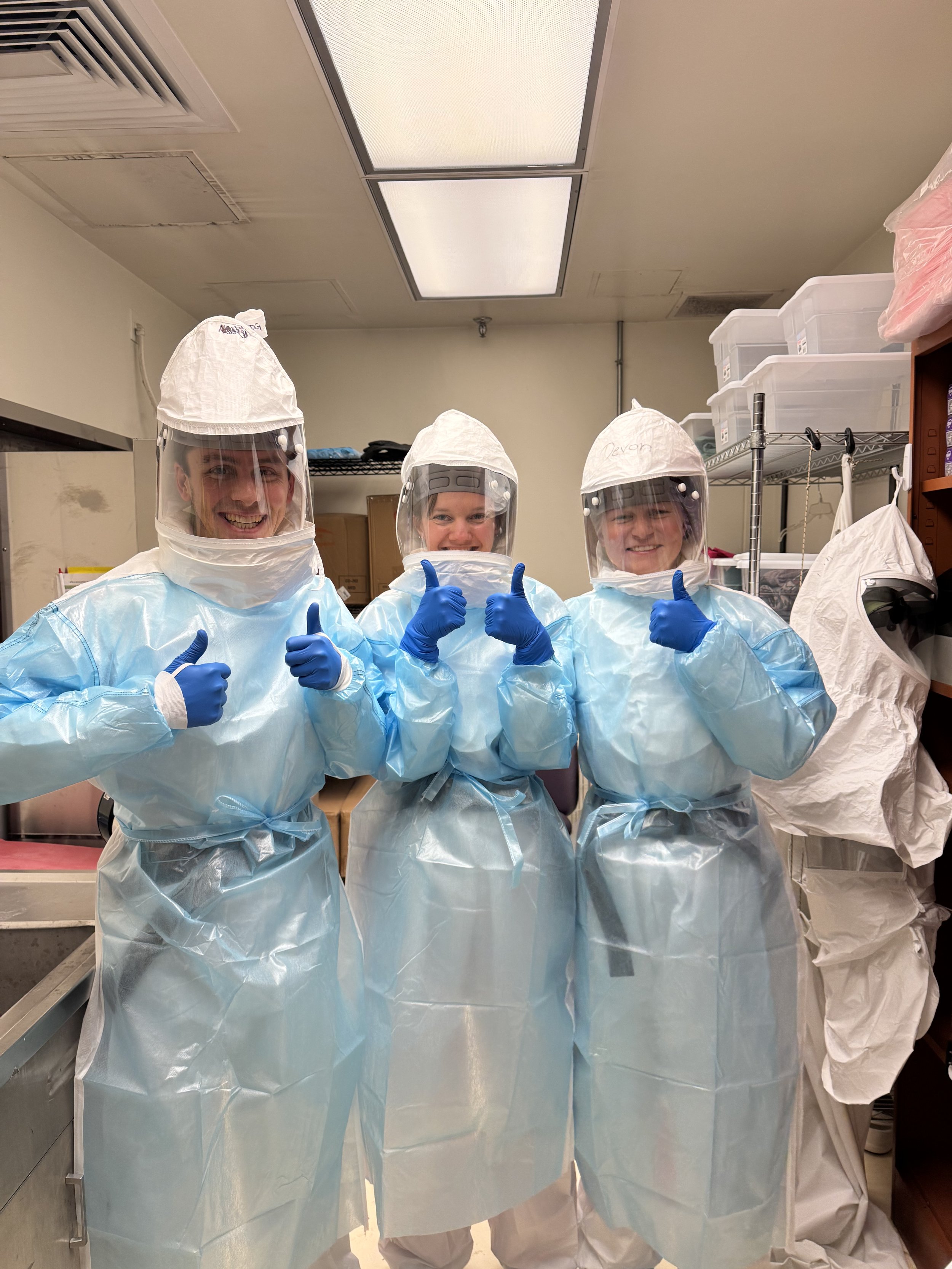 Three healthcare workers wearing full personal protective equipment, including face shields, gloves, and gowns, standing in a medical room and giving thumbs up.