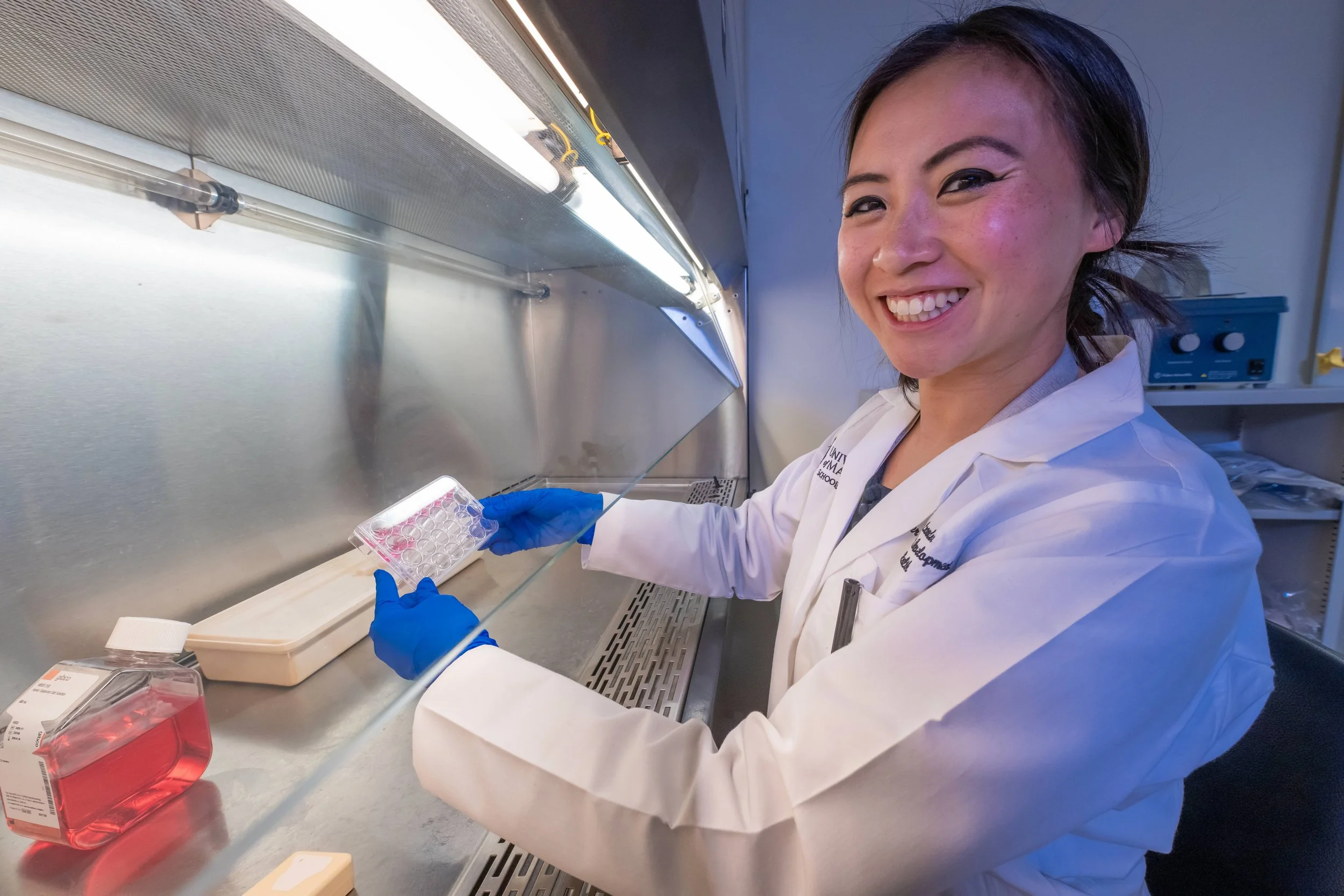 A scientist working in a laboratory inside a biosafety cabinet, wearing a white lab coat and blue gloves, holding a test tube rack while smiling at the camera.