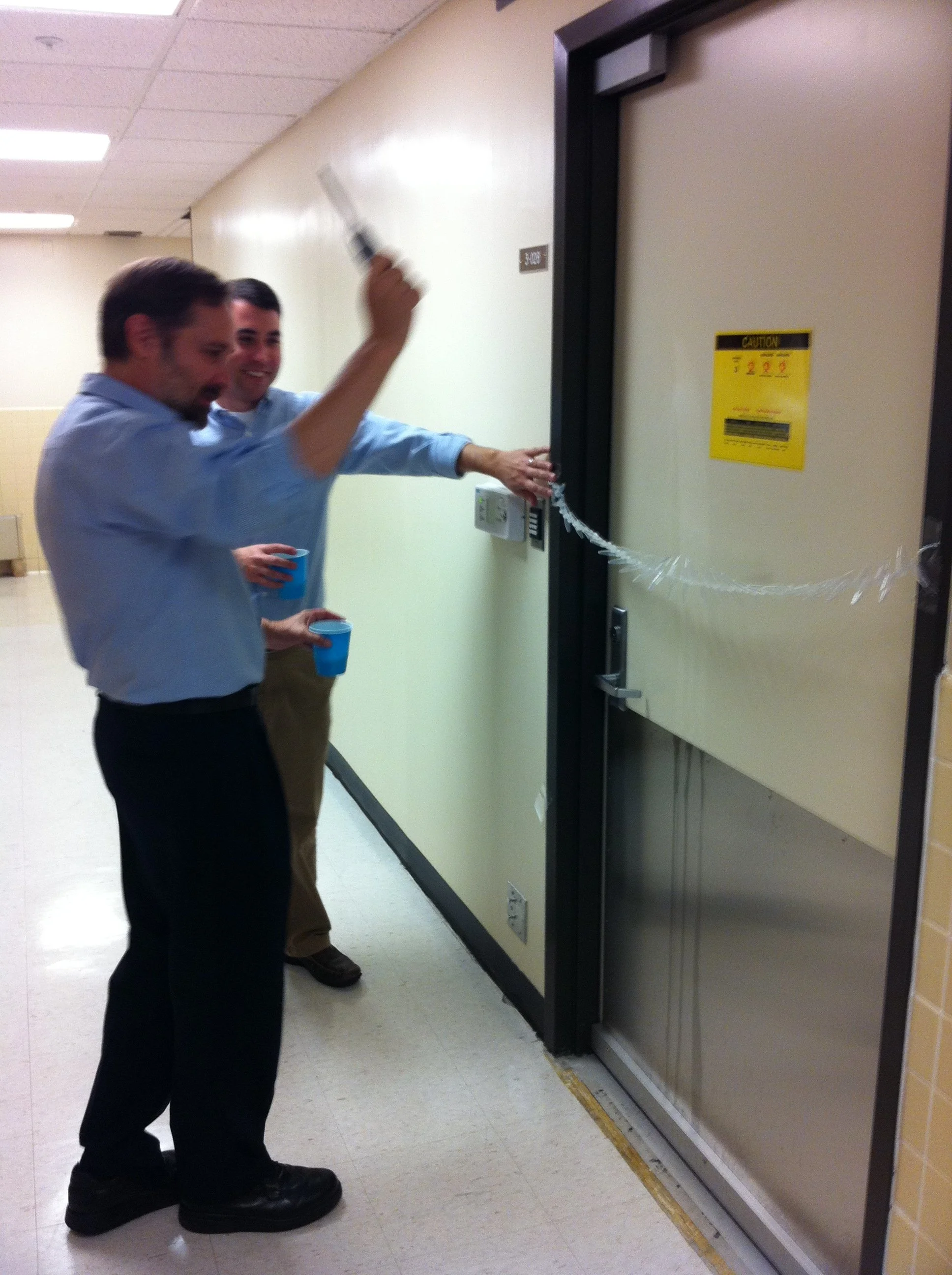 Two men in office attire celebrating after breaking a plastic retractable door safety strip, with drinks in their hands, in a hallway.