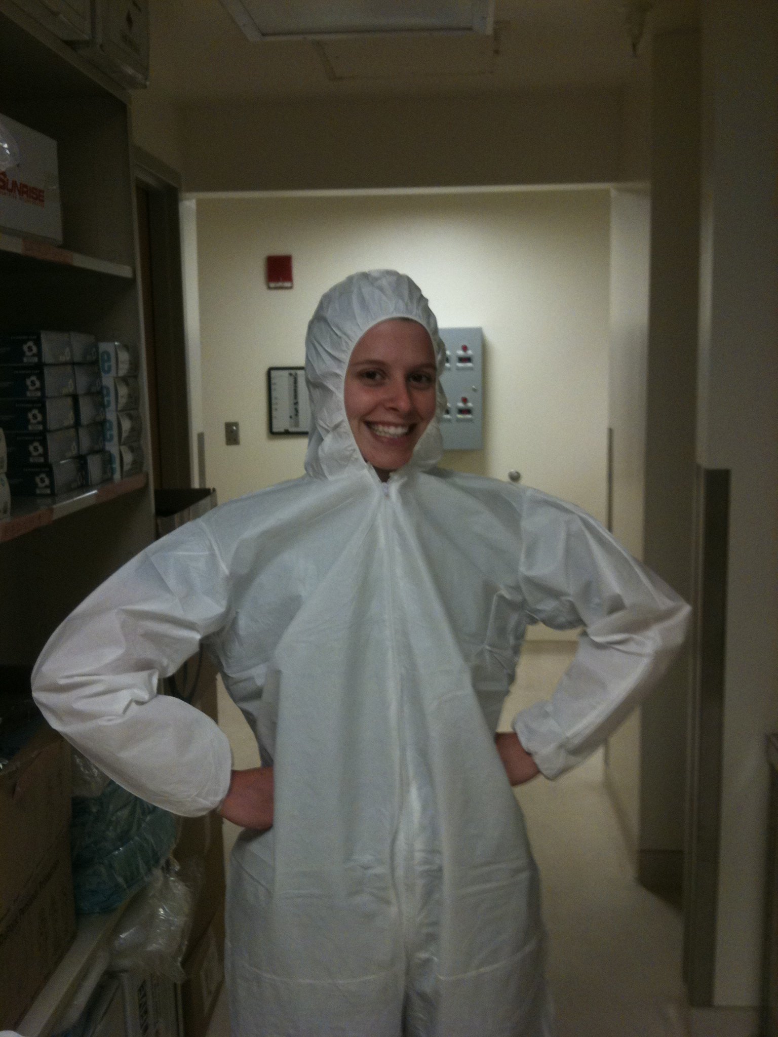 A woman in a white protective coverall suit standing in a hospital or laboratory hallway, smiling with hands on hips.
