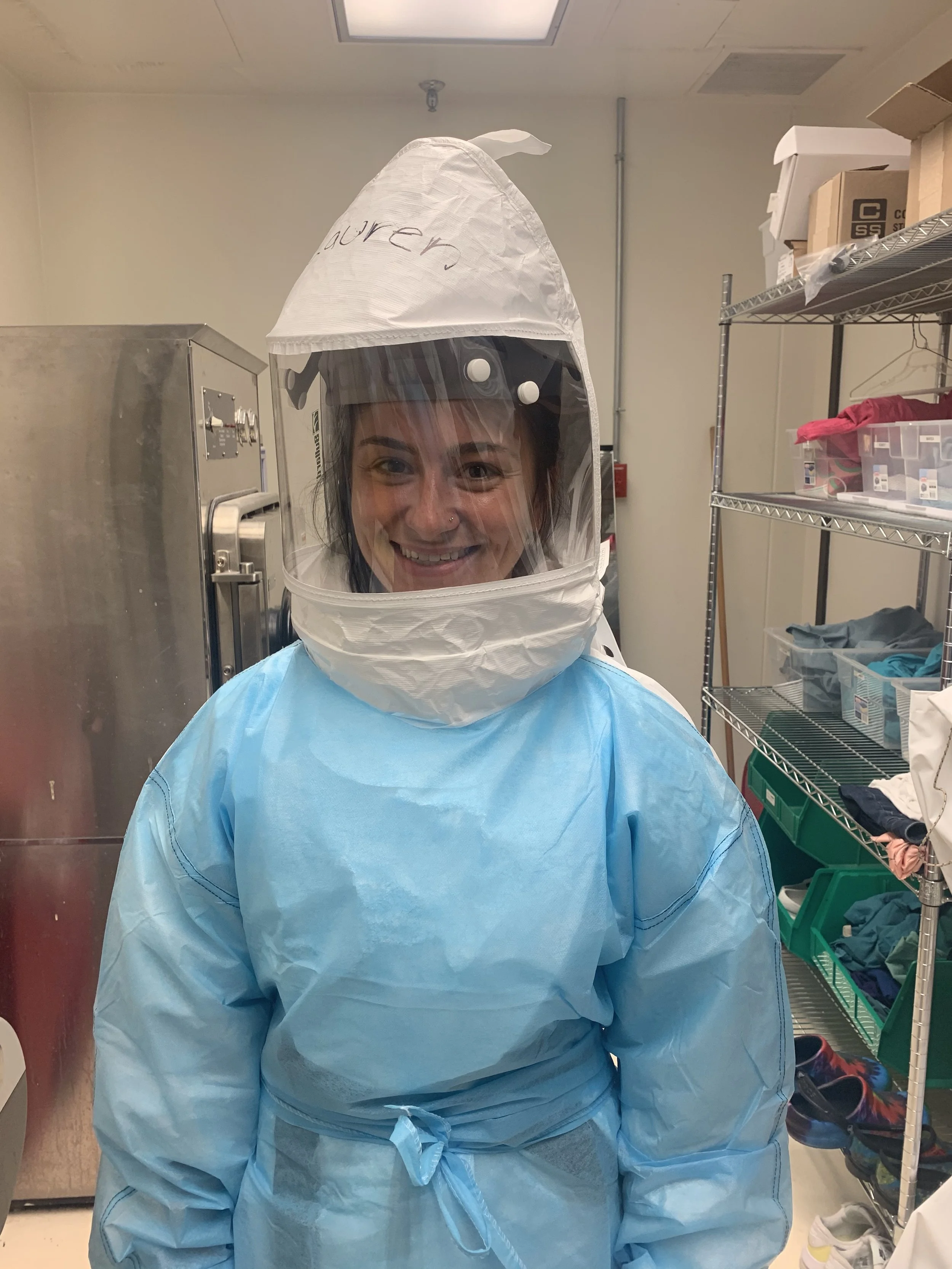 A woman dressed in full medical PPE, including a light blue gown and a protective hood with a clear face shield. She is smiling and standing in a medical or laboratory setting with shelves and equipment around her.
