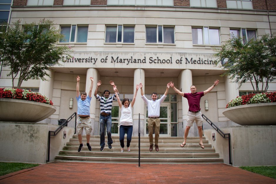 A group of five people jumping in front of the University of Maryland School of Medicine building.
