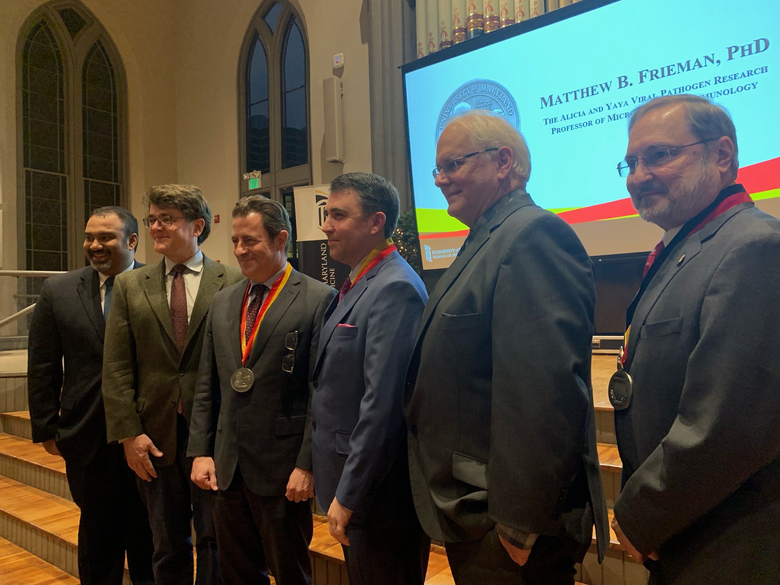 Six men in formal suits standing in a row on stage, some wearing medals, in front of a large screen. The screen displays the name Matthew B. Frieman, PhD, and his affiliation with the University of Maryland.