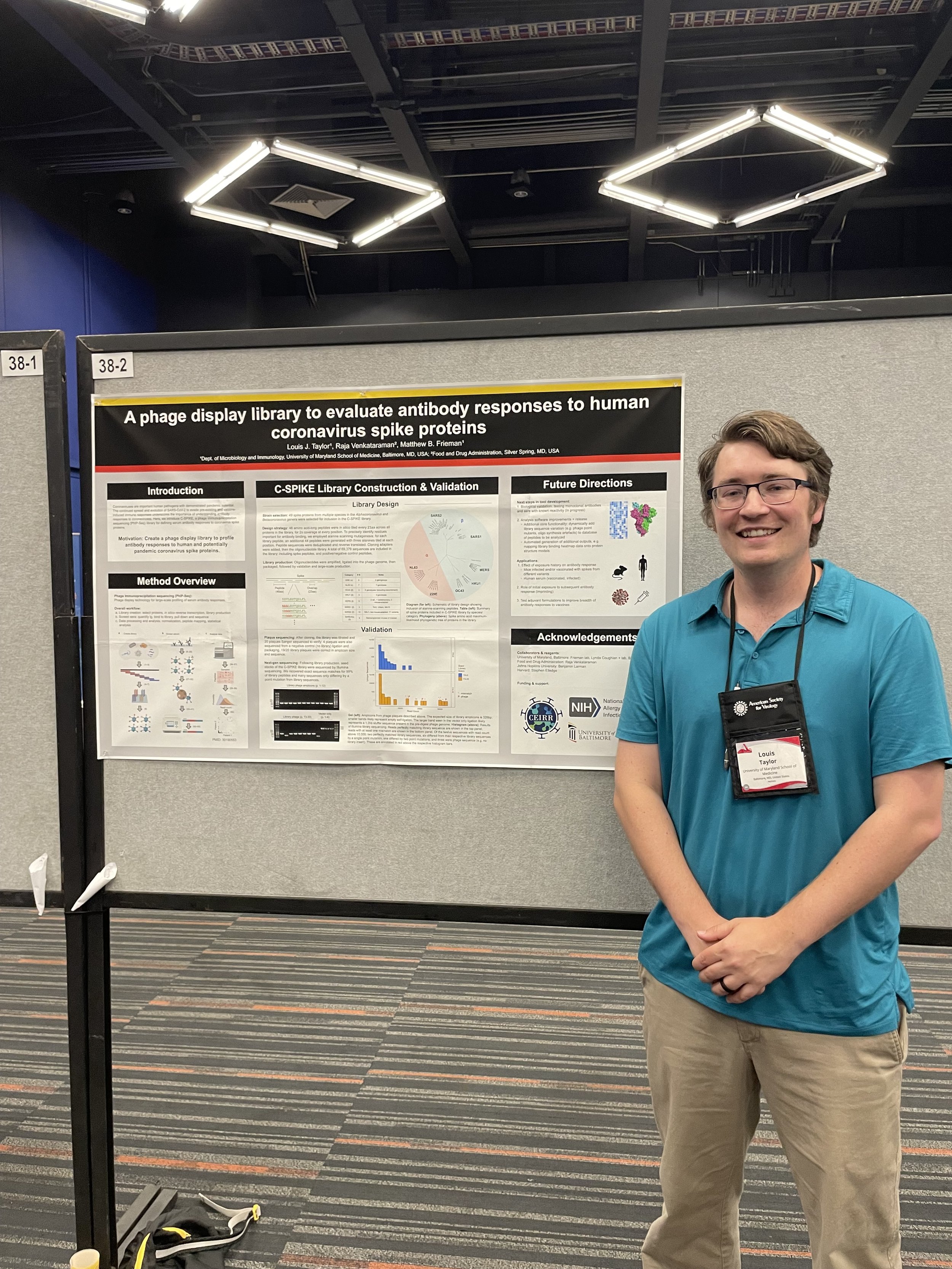 A man in a blue shirt and glasses standing in front of a scientific research poster at a conference. The poster discusses a phage display library for evaluating antibody responses to human coronavirus spike proteins.